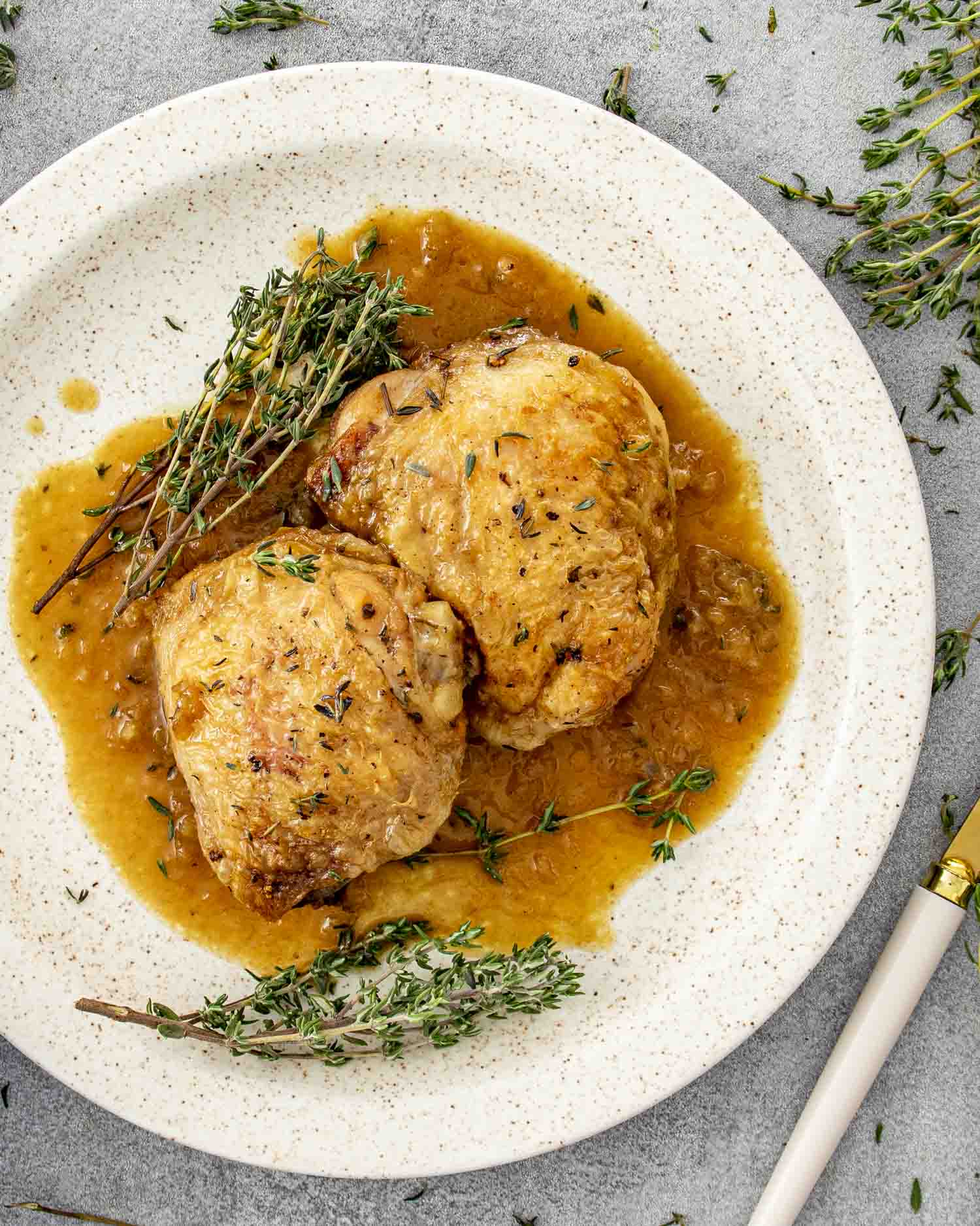 Two apple cider glazed chicken thighs on a plate with thyme and plenty of sweet-tangy sauce pooling underneath.