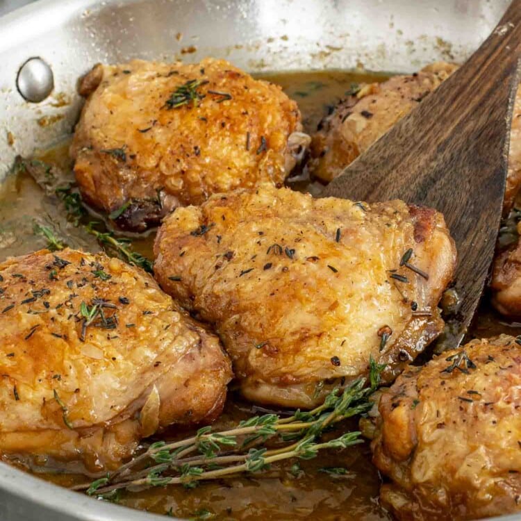 Crispy chicken thighs simmering in a rich apple cider sauce with thyme sprigs, cooked in a stainless-steel skillet.