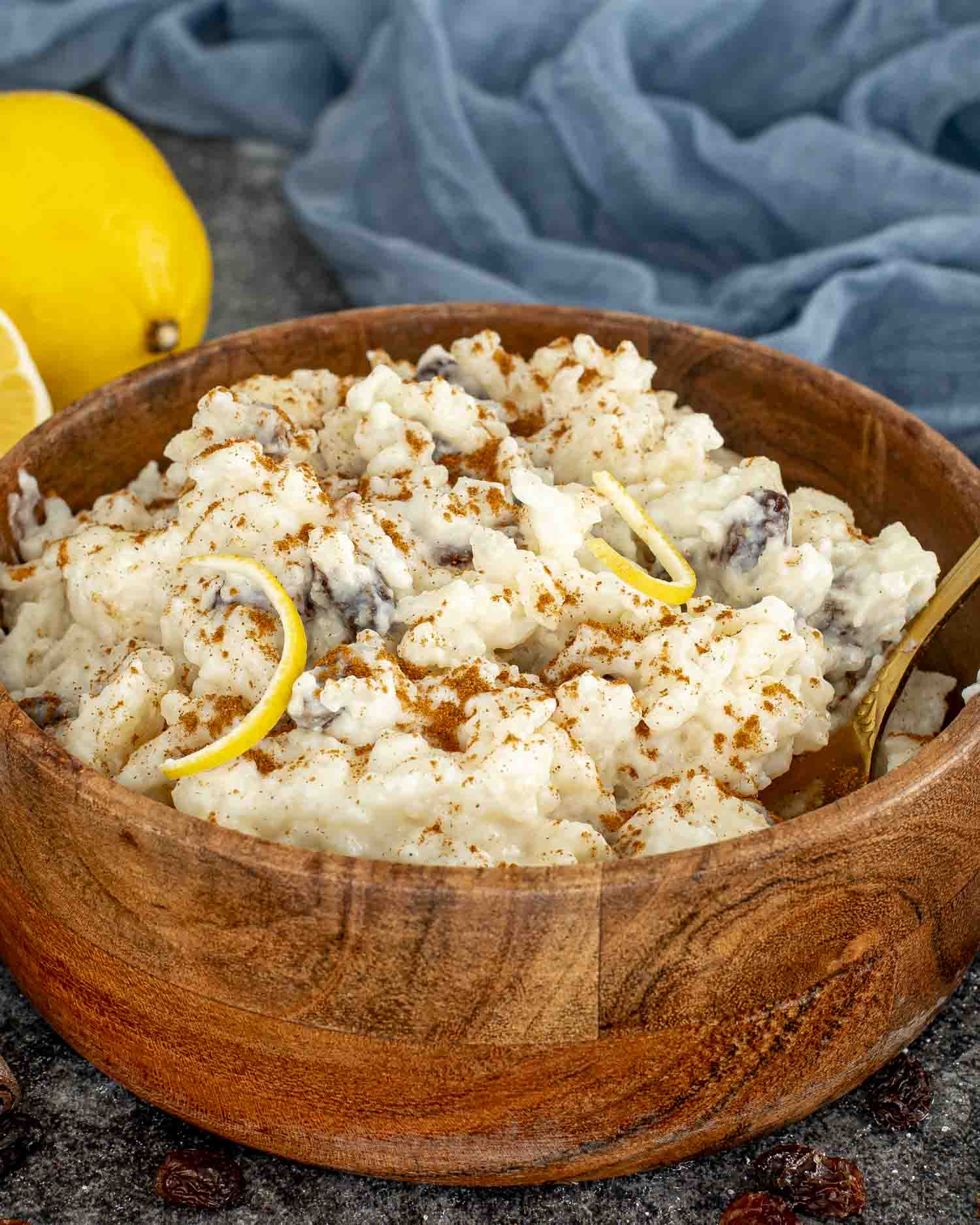 Wooden bowl with creamy arroz con leche, raisins, cinnamon, and lemon zest, set on dark surface.