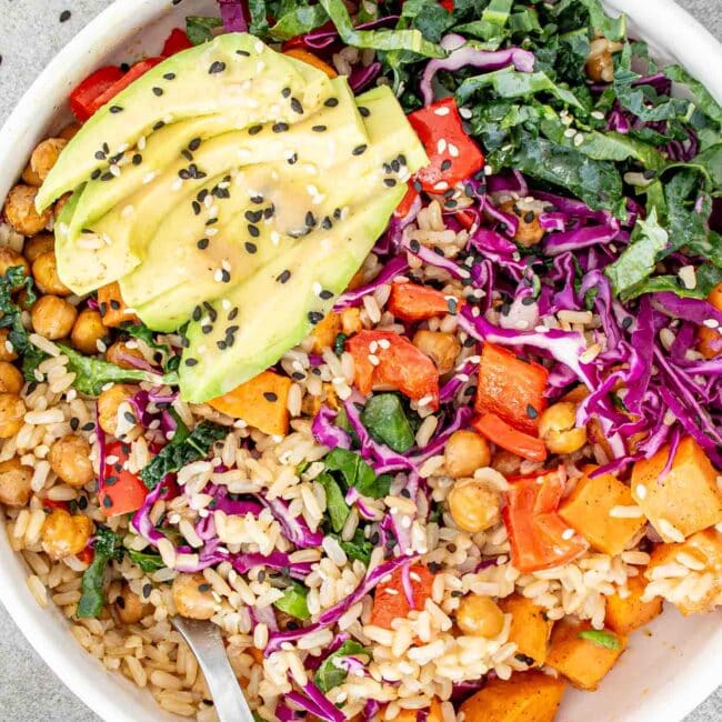 A colorful Buddha bowl with avocado, chickpeas, rice, and mixed vegetables, garnished with black sesame seeds.