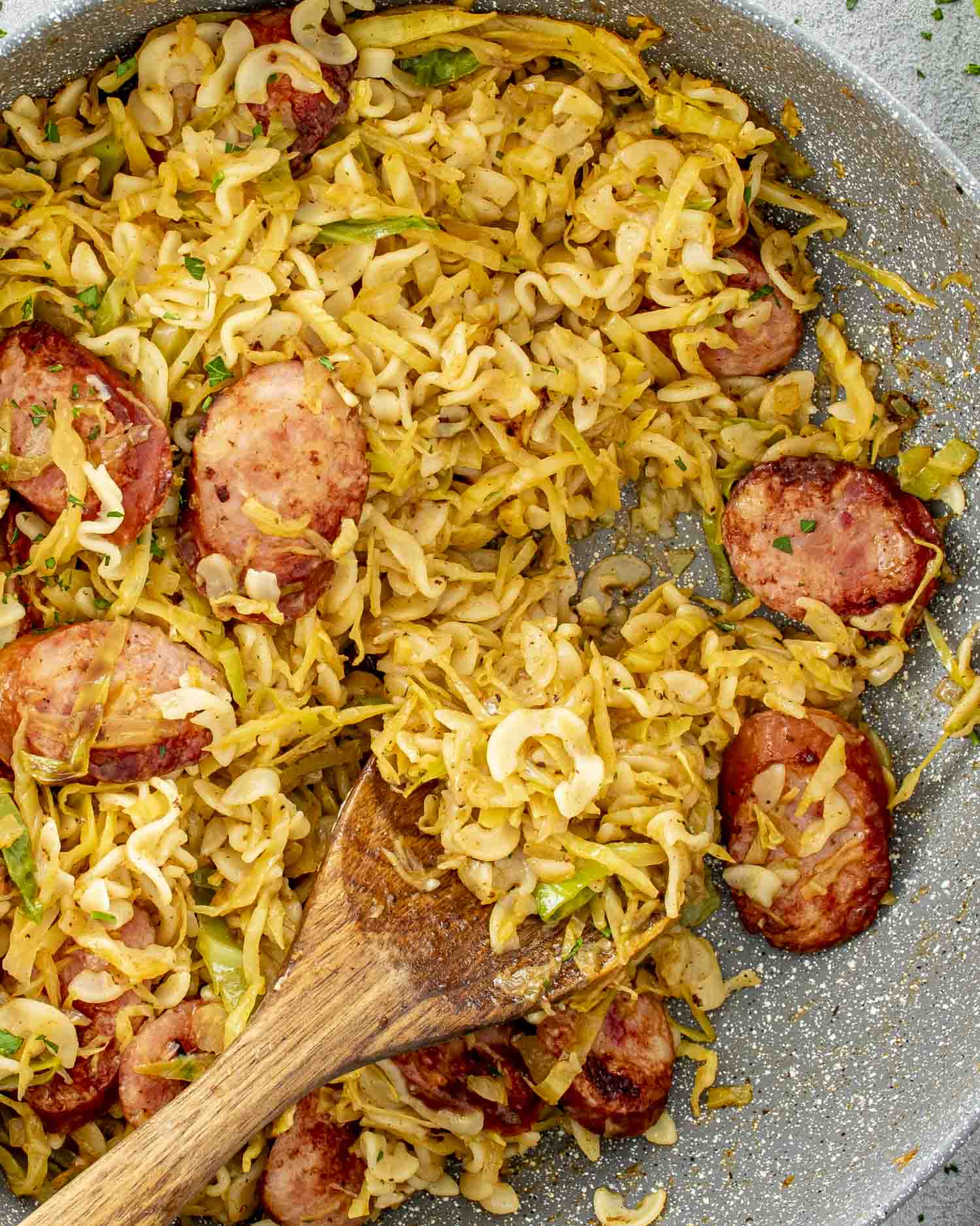 a big skillet with freshly made cabbage and noodles with sausage with a wooden spoon inside.