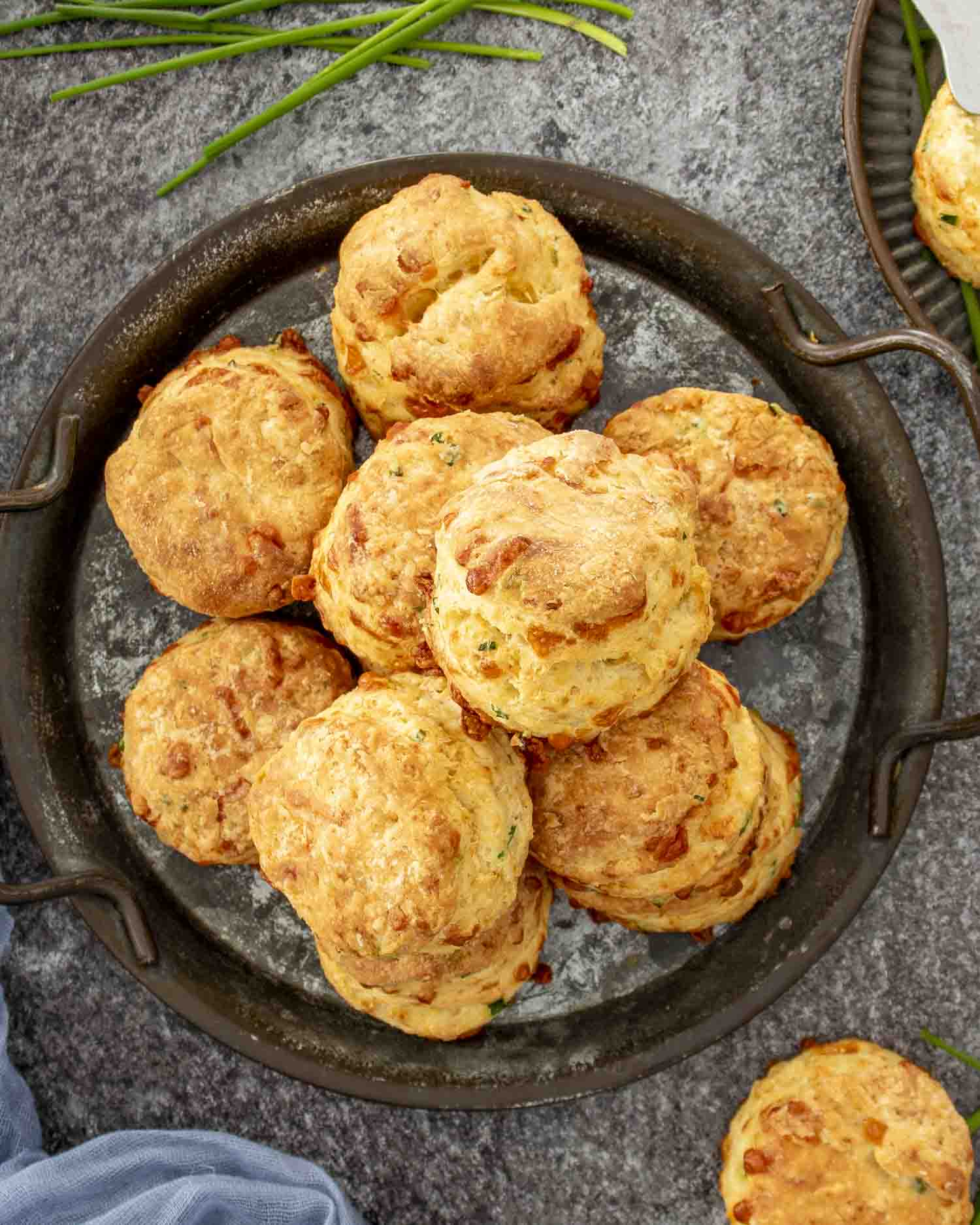A plate piled with cheddar chive biscuits, surrounded by fresh chives and a rustic surface underneath.