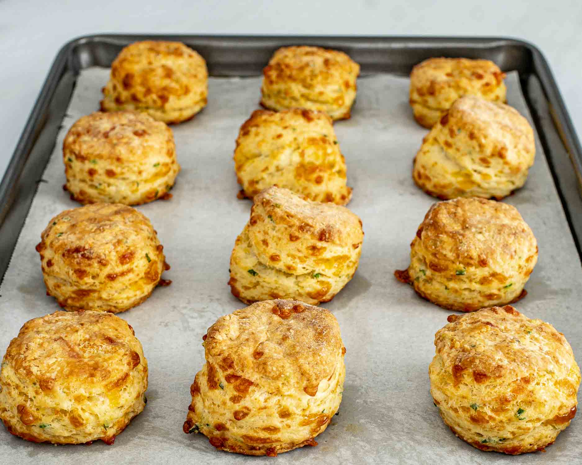 Golden cheddar chive biscuits just out of the oven on a parchment-lined baking sheet, showing tall flaky layers.