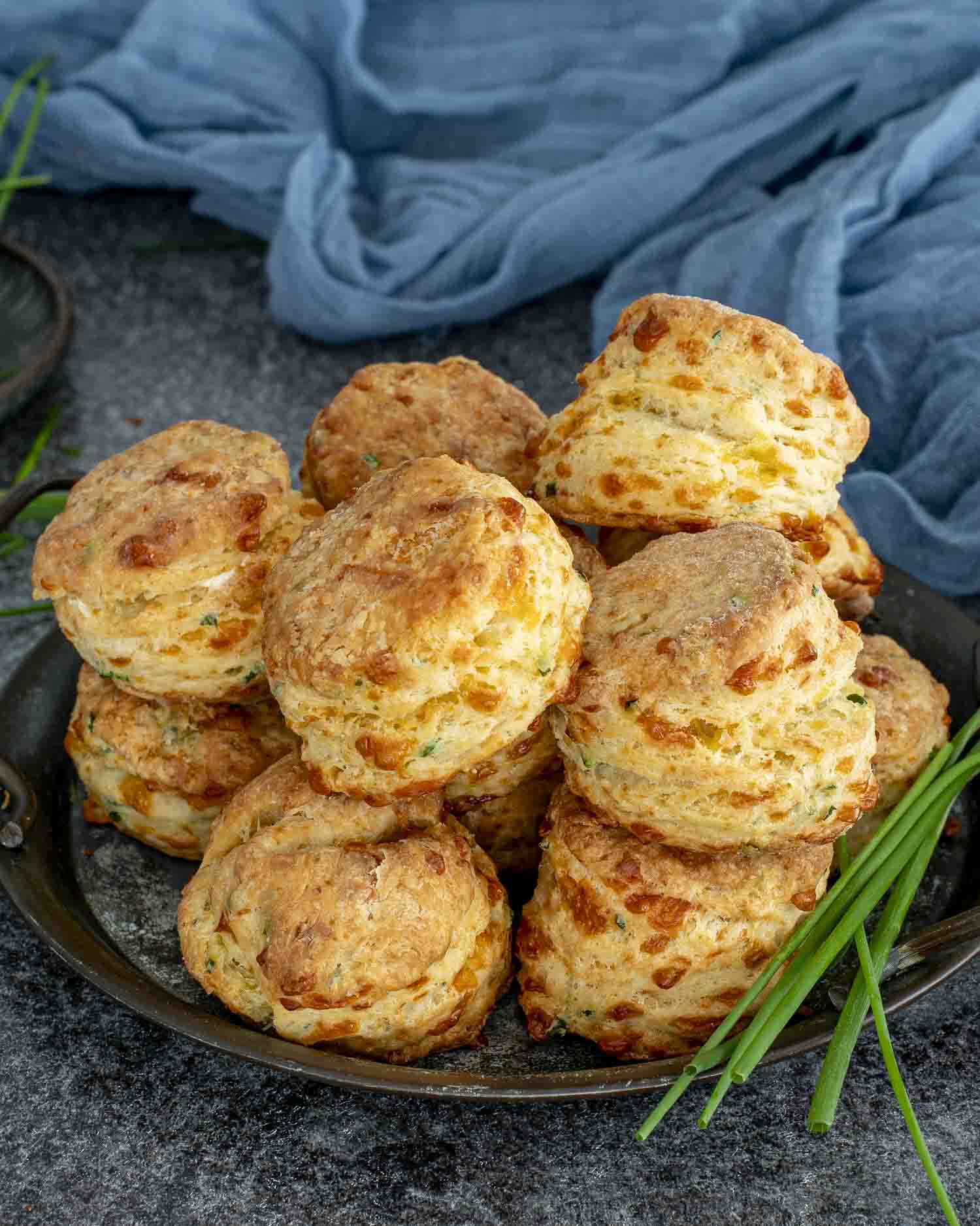 Freshly baked cheddar chive biscuits stacked high on a dark tray, showing golden flaky layers and melted cheddar throughout.