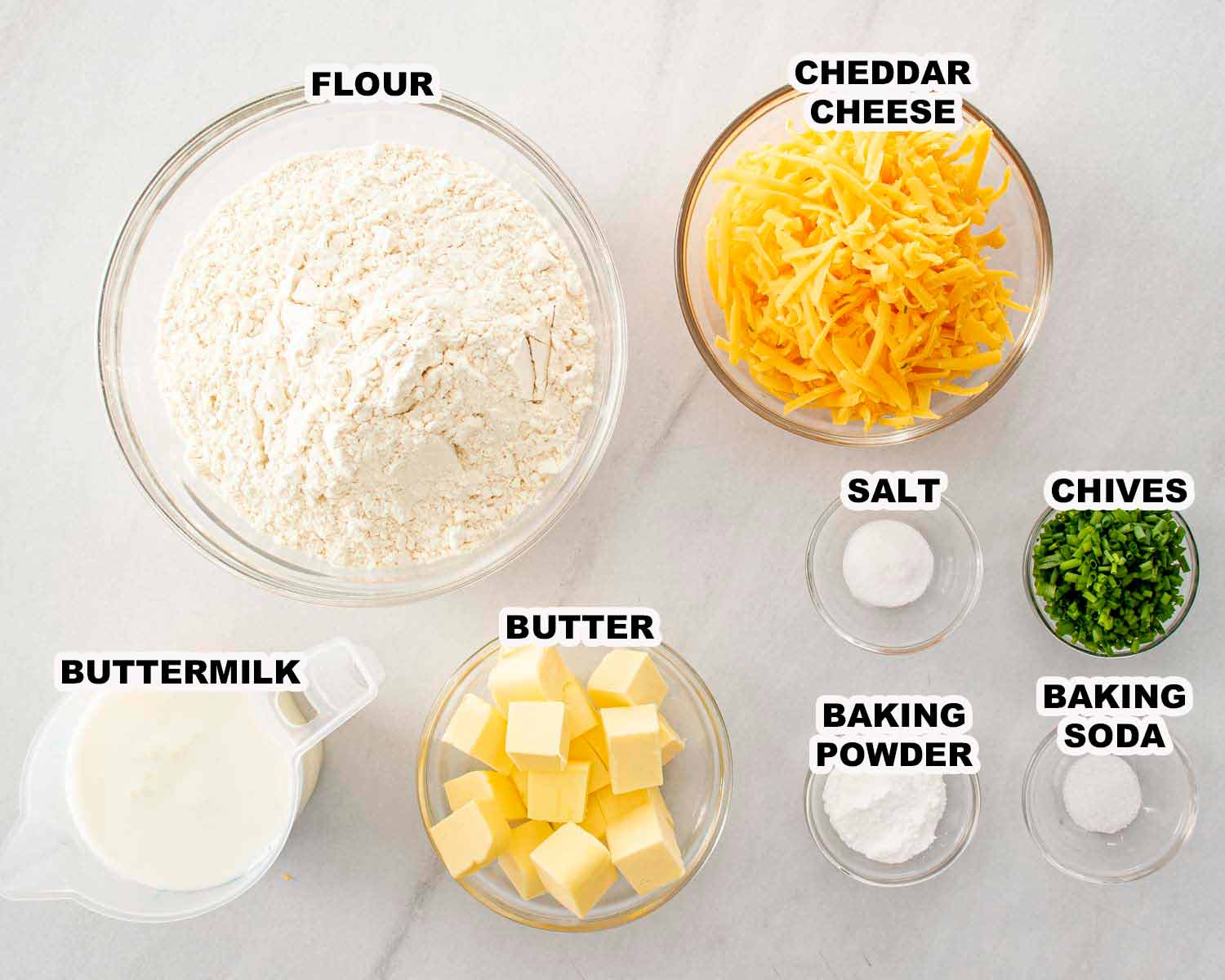 Flour, cheddar cheese, butter, buttermilk, chives, baking powder, baking soda, and salt arranged in bowls for cheddar chive biscuits.