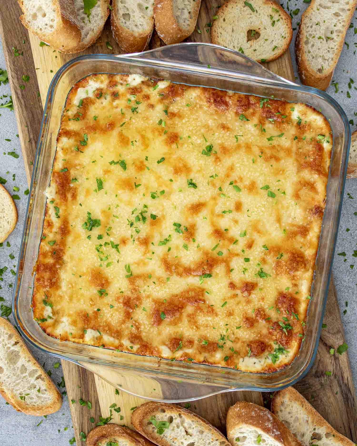 Golden, bubbly cheesy roasted garlic dip baked in a glass dish, topped with parsley, served with crusty baguette slices.