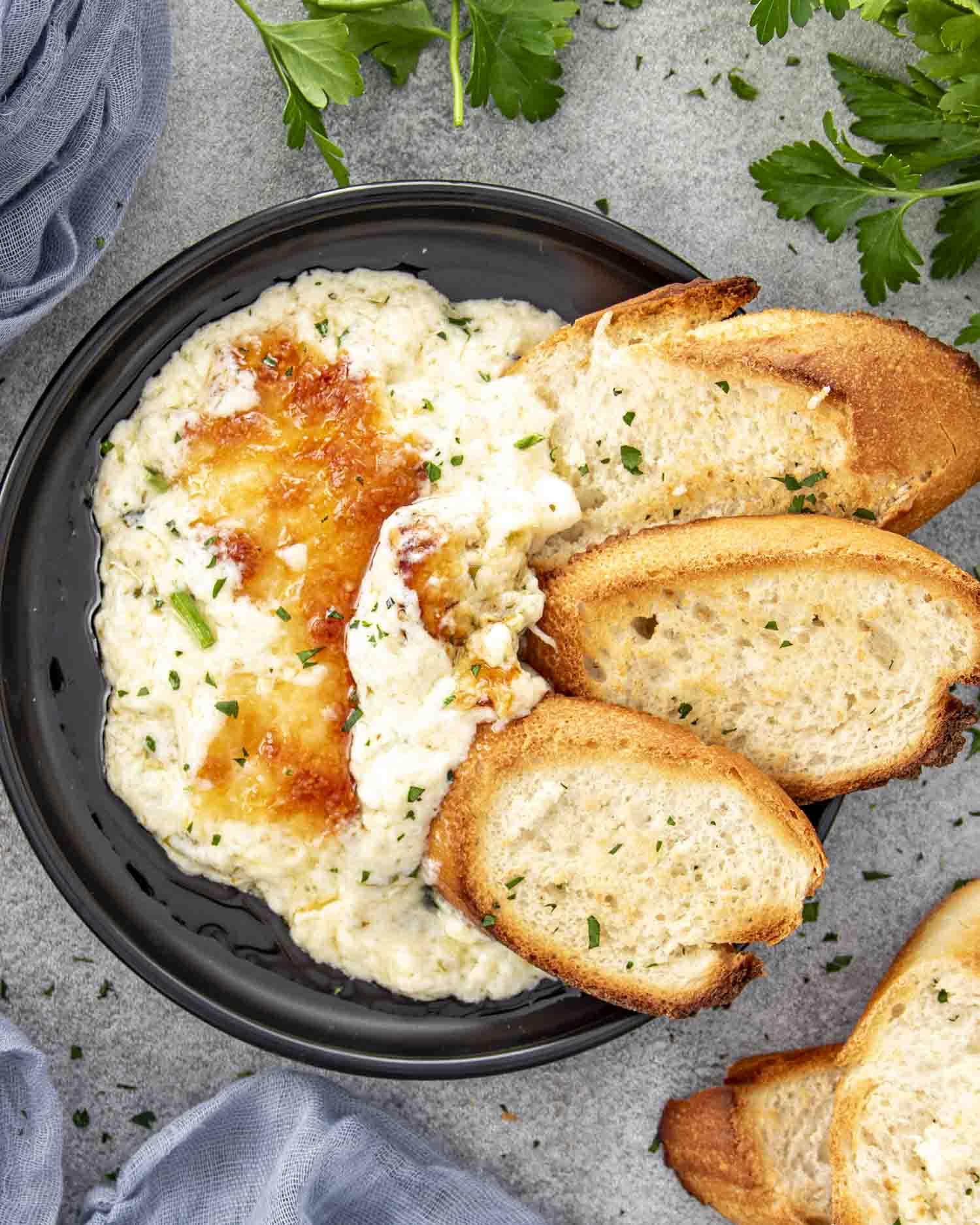 Close-up of creamy roasted garlic dip in a black bowl, topped with melted cheese, served with toasted baguette slices.