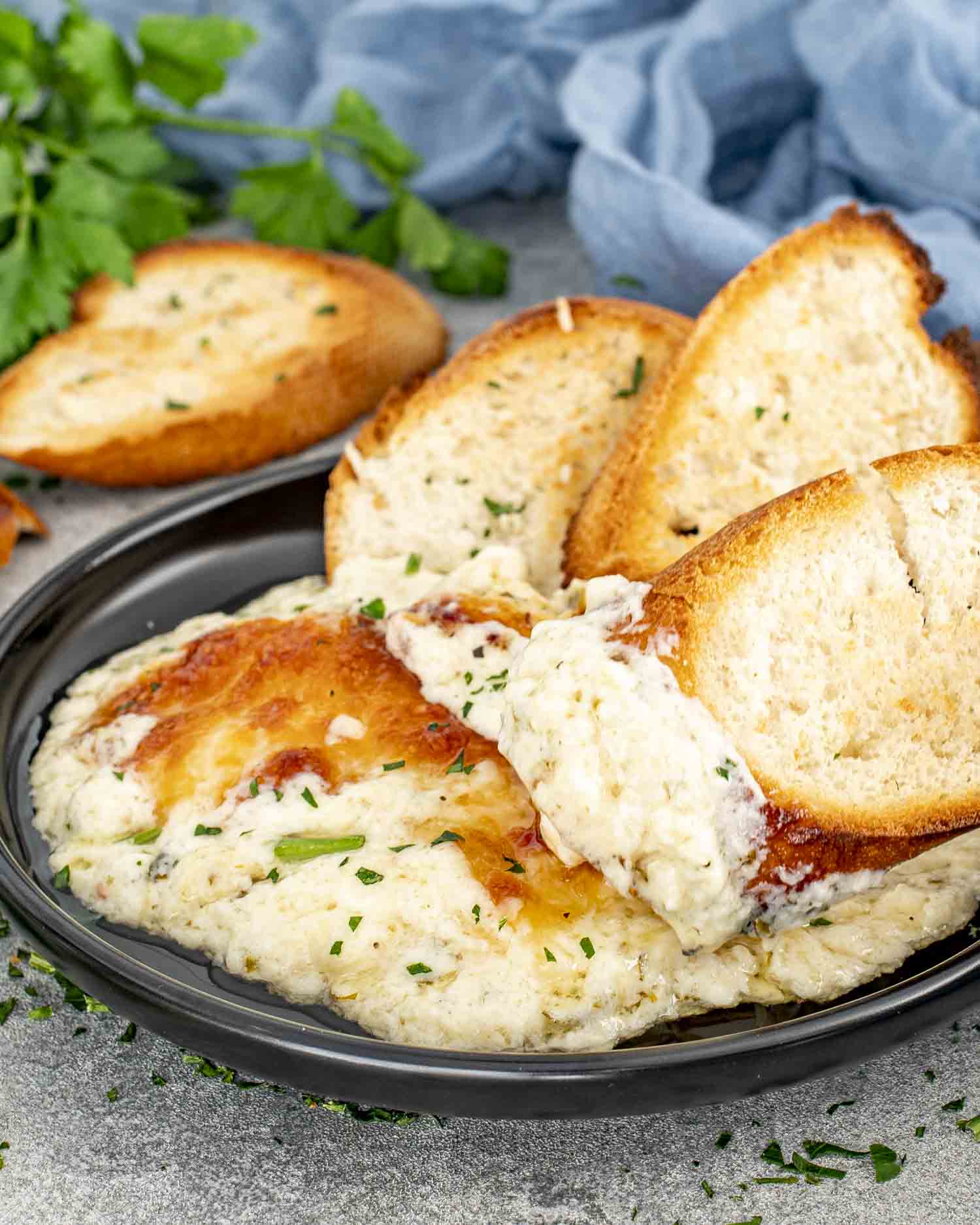 Creamy roasted garlic dip served in a black bowl, topped with golden cheese, surrounded by toasted baguette slices for dipping.