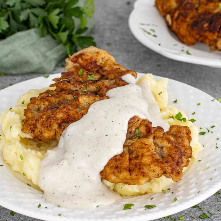 Plate of golden chicken fried steak with gravy over mashed potatoes, garnished with parsley, fork, and gravy boat nearby.