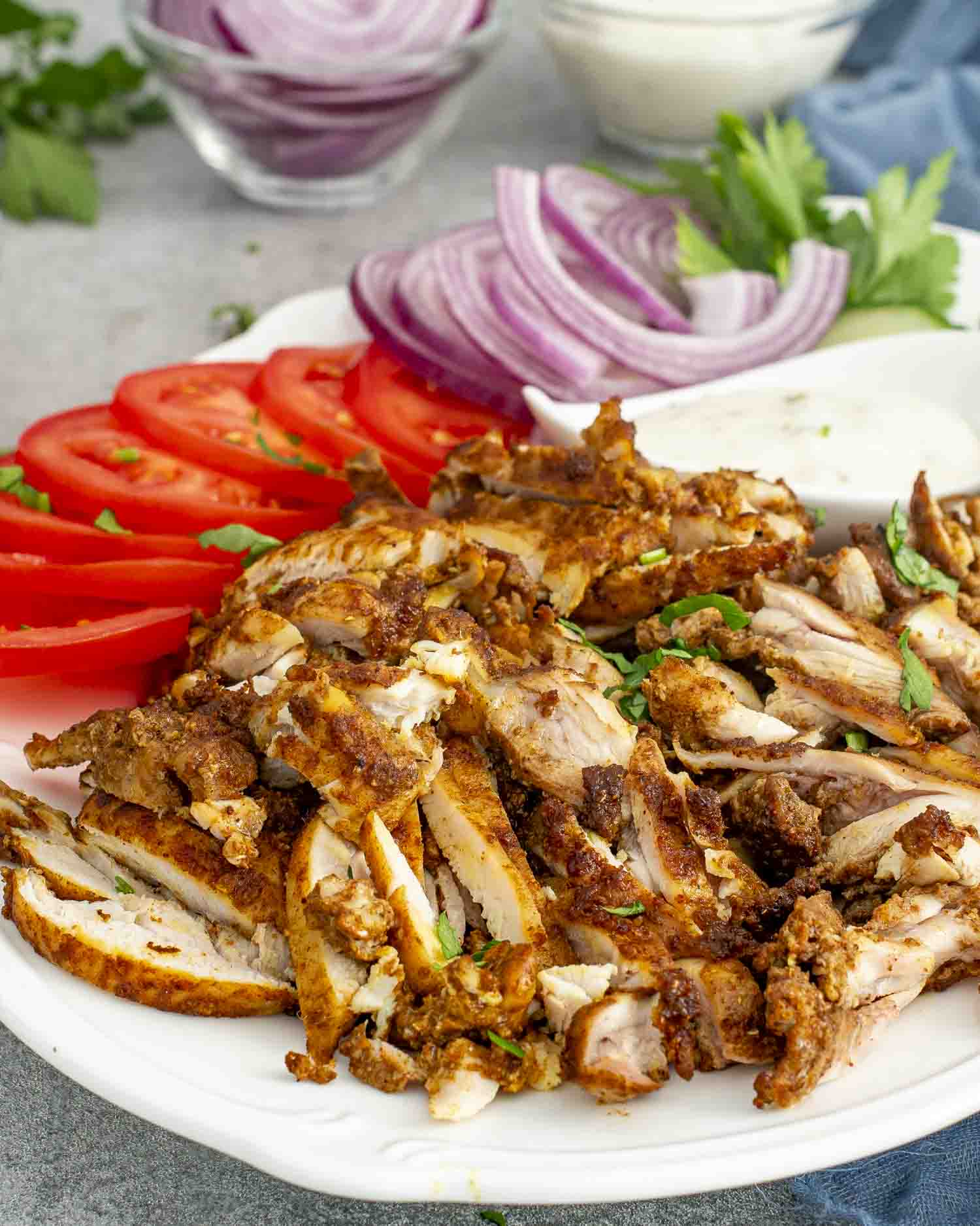 A plate of sliced Chicken Shawarma, served with fresh tomato slices, red onion, cucumber, parsley, and a creamy tzatziki sauce.
