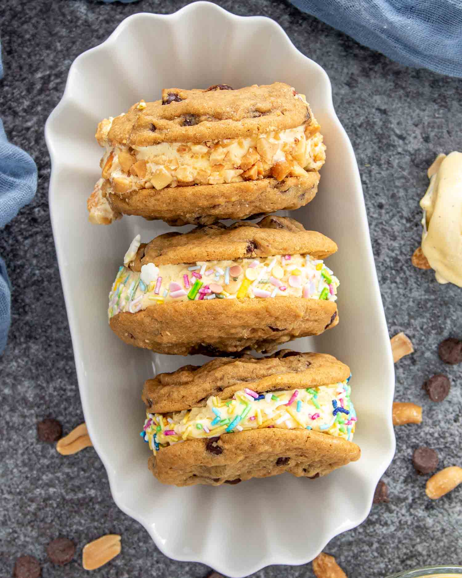 Top view of chocolate chip cookie ice cream sandwiches in a scalloped white dish, with colorful sprinkles and chopped peanuts.