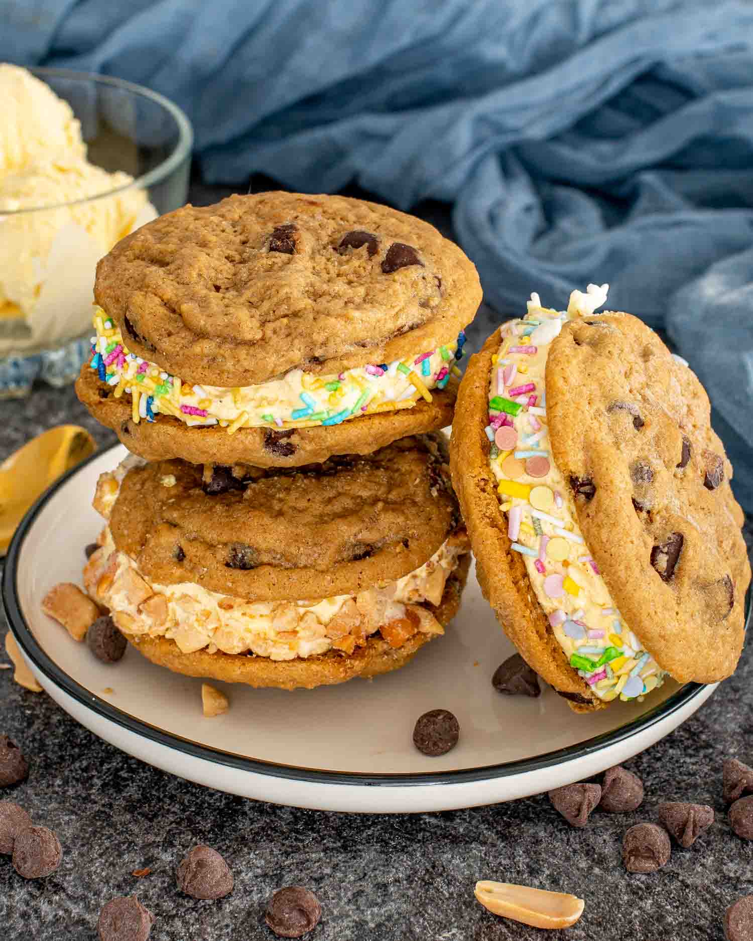 Three chocolate chip cookie ice cream sandwiches on a plate, each rolled in colorful sprinkles or chopped peanuts.