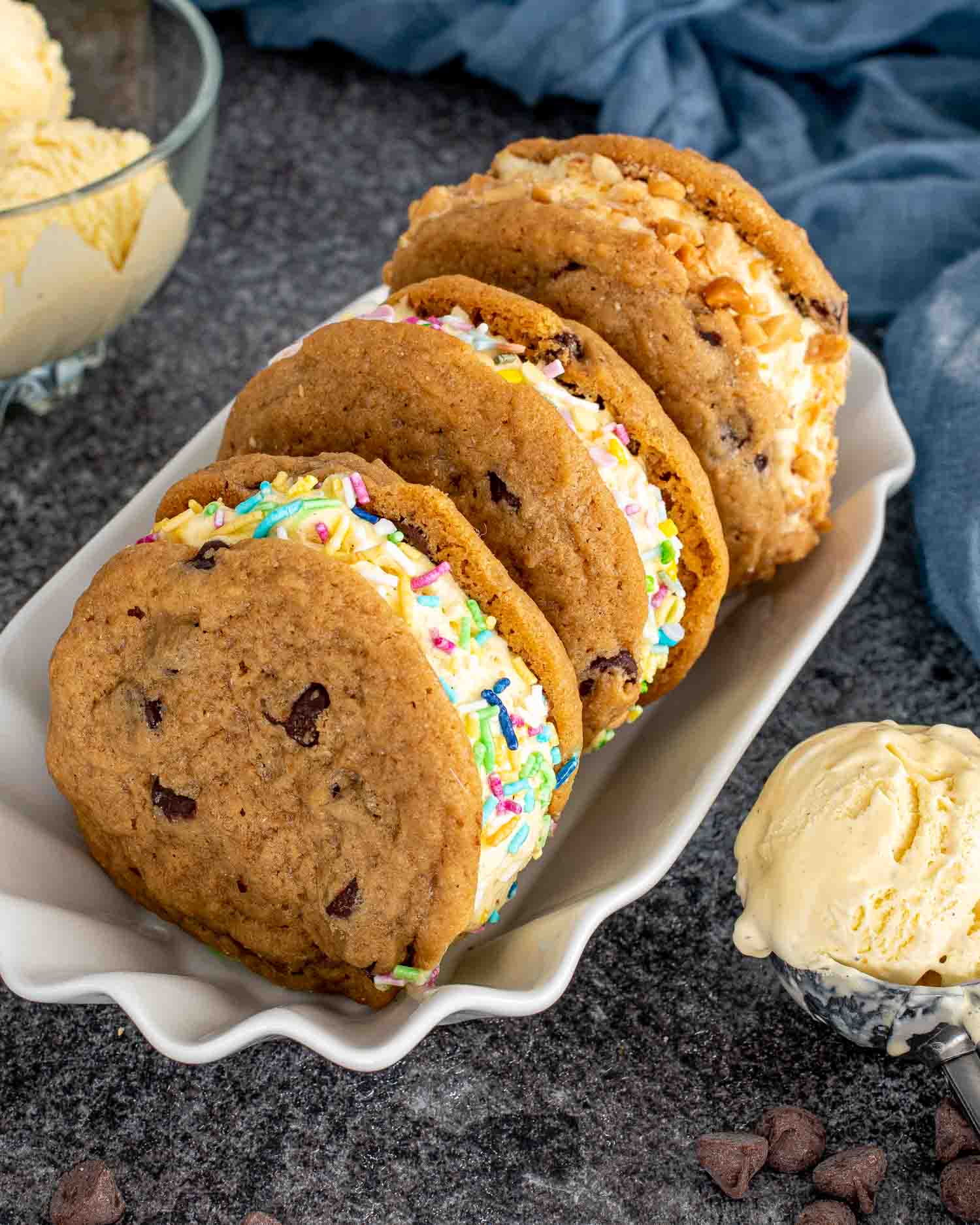 Row of chocolate chip cookie ice cream sandwiches in a white dish, rolled in sprinkles and chopped peanuts, with ice cream scoop.