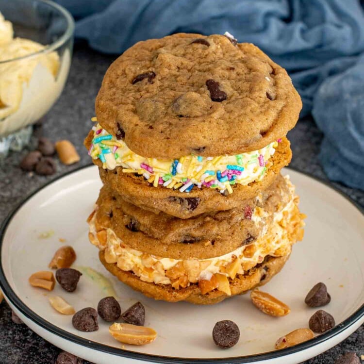 Stack of chocolate chip cookie ice cream sandwiches with colorful sprinkles and chopped peanuts, chocolate chips scattered on the table.