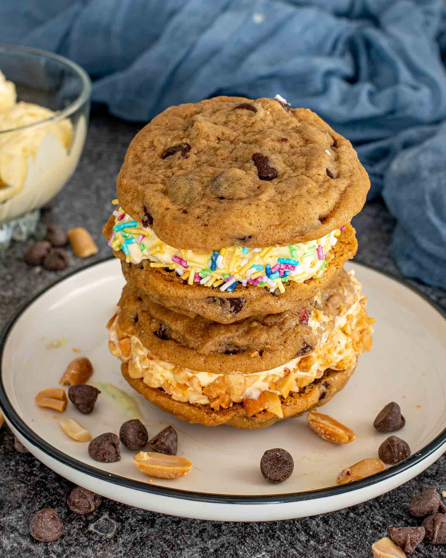 Stack of chocolate chip cookie ice cream sandwiches with colorful sprinkles and chopped peanuts, chocolate chips scattered on the table.