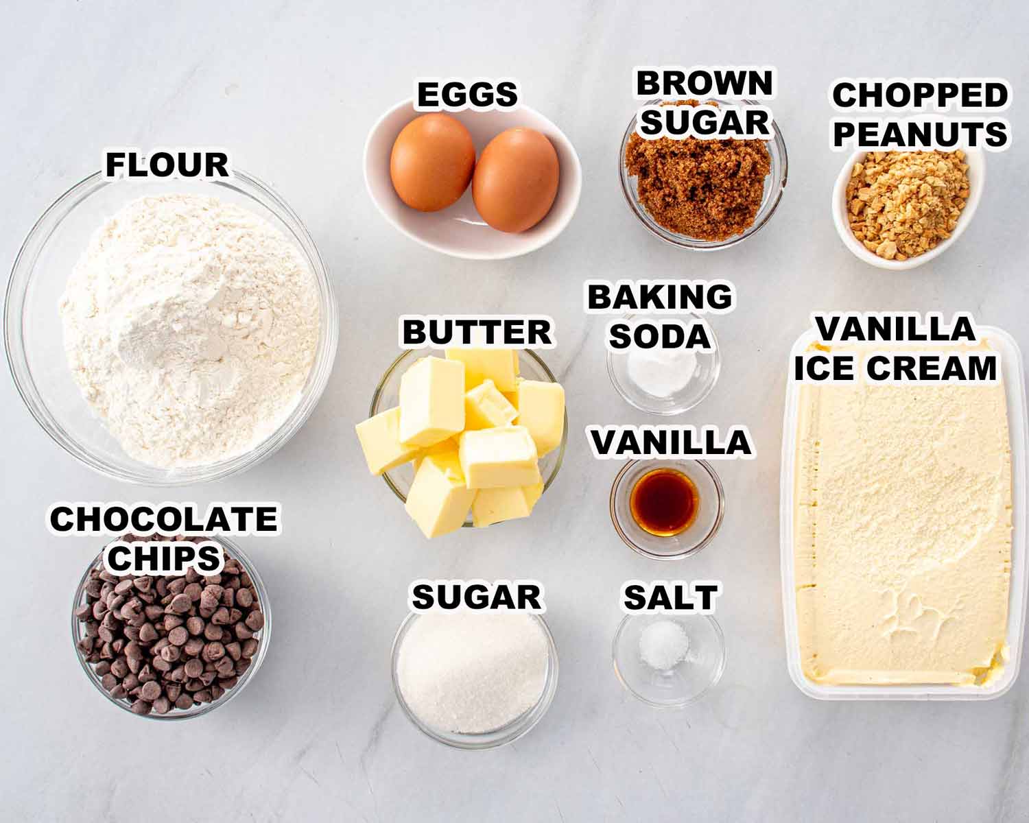 Overhead view of labeled ingredients for chocolate chip cookie ice cream sandwiches including flour, butter, sugar, eggs, and vanilla ice cream.