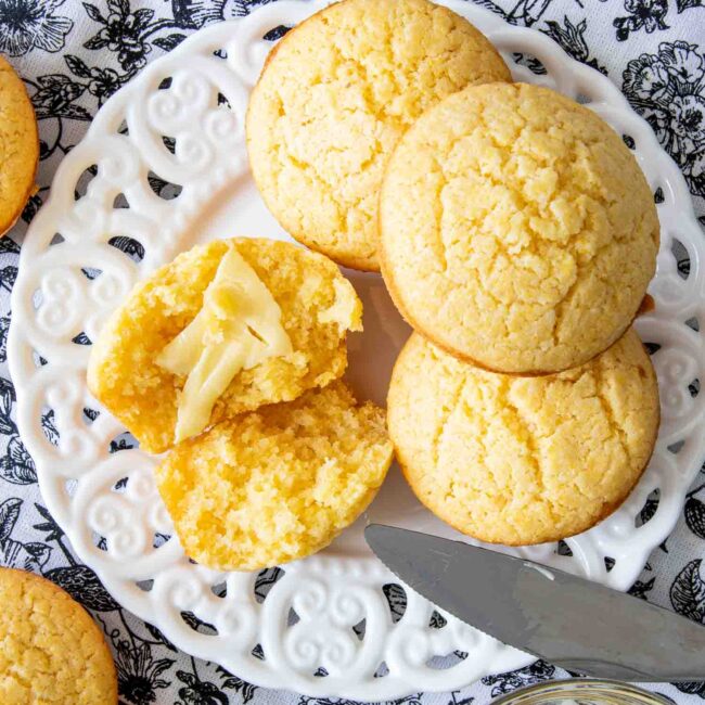 a cornbread muffin broken in half with butter on a plate with a few other cornbread muffins.