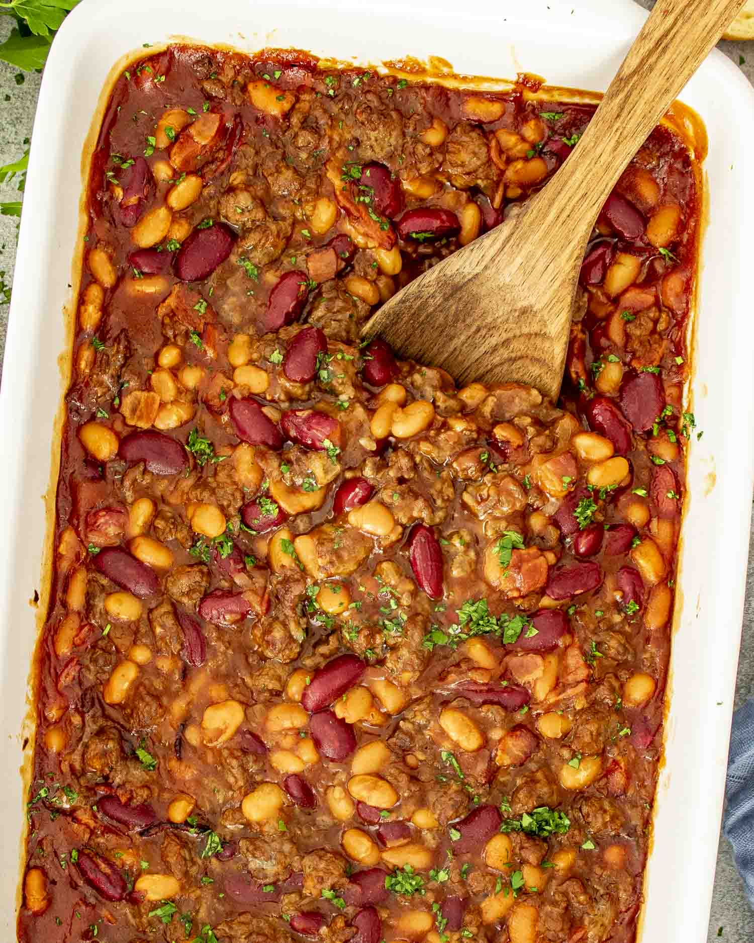 Large baking dish full of cowboy baked beans, with wooden spoon scooping from one side.