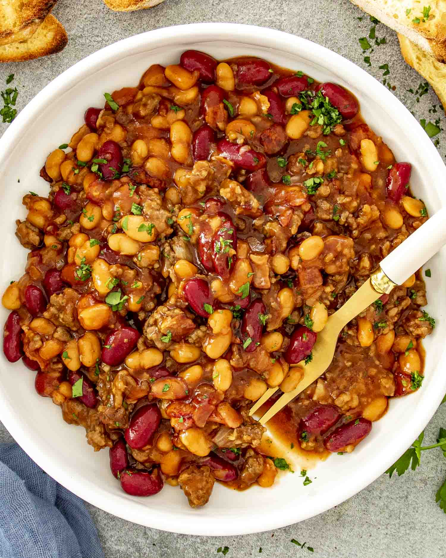 Top-down view of a white bowl filled with meaty baked beans, served with a gold fork.