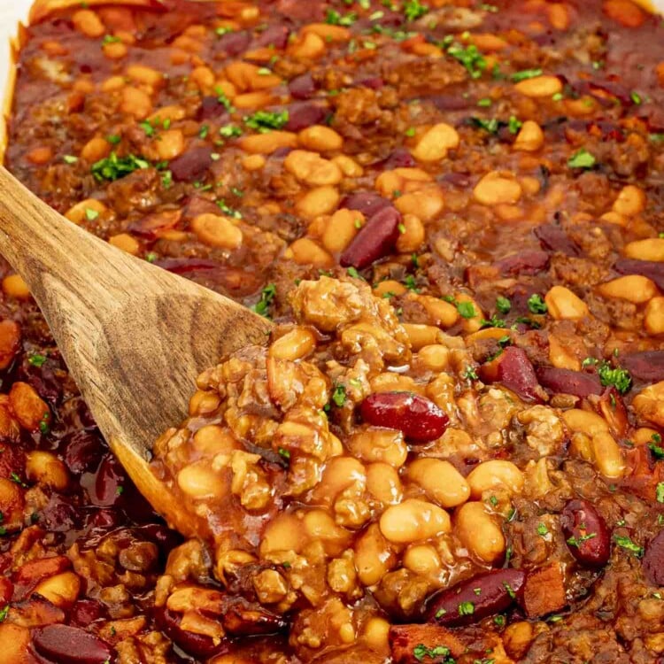 Casserole dish filled with baked beans, bacon, and beef, being scooped with a wooden spoon.