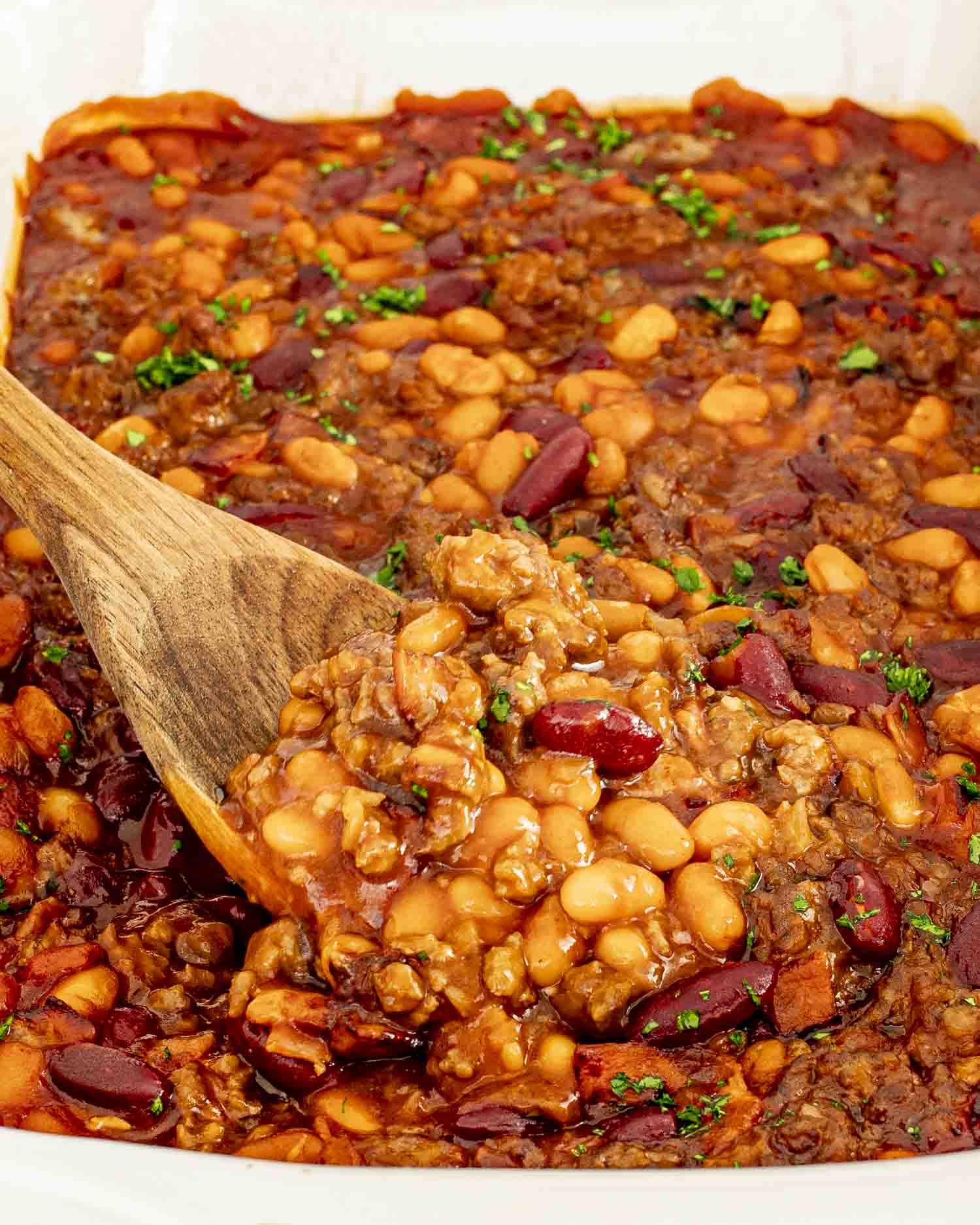 Casserole dish filled with baked beans, bacon, and beef, being scooped with a wooden spoon.