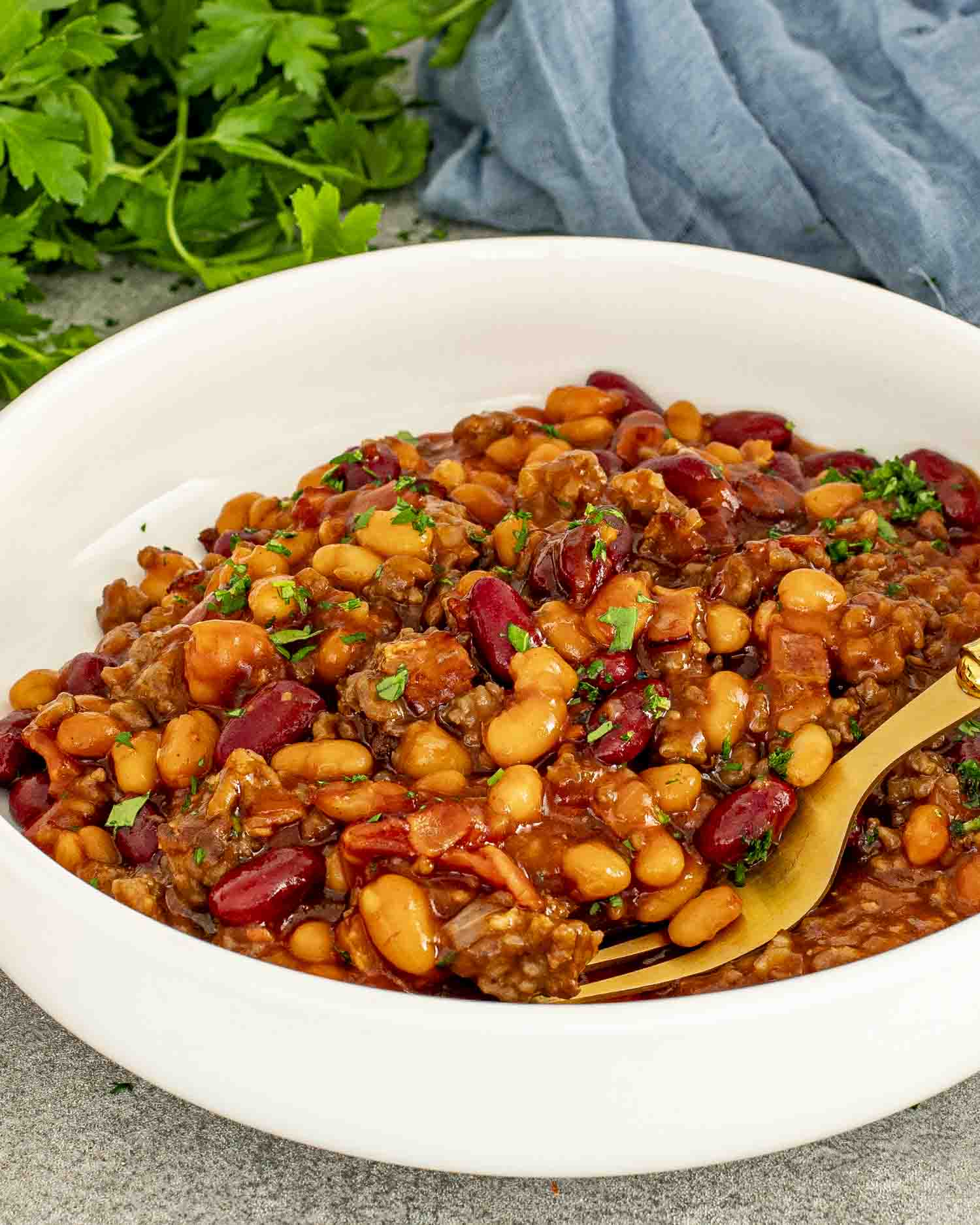 White bowl of cowboy baked beans with bacon, beef, and two types of beans, garnished with parsley.