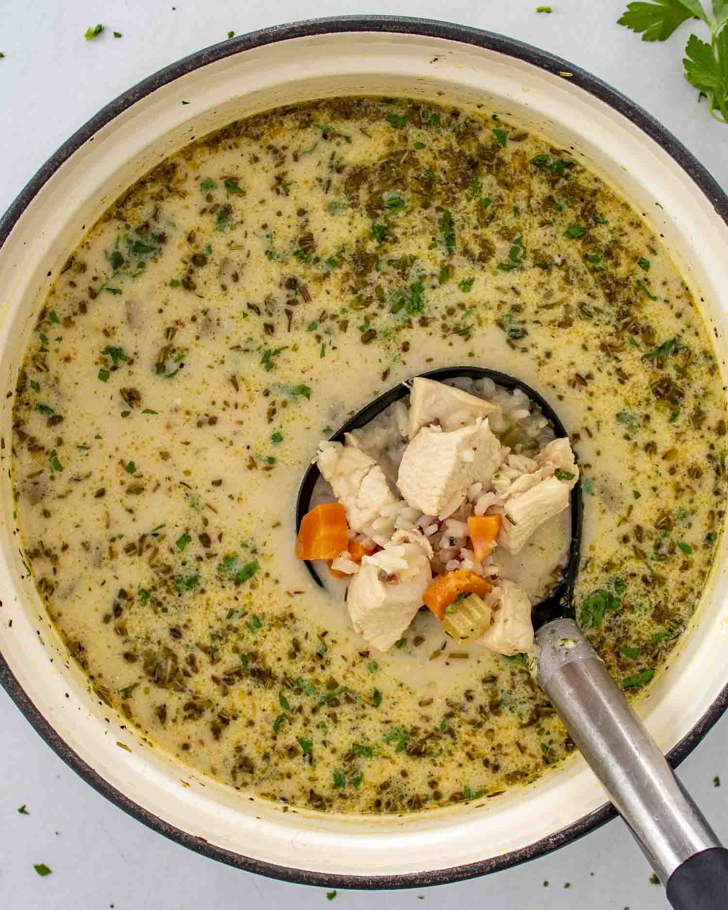 A close-up of a creamy chicken wild rice soup in a large enameled pot. A black ladle is scooping up a portion, showing tender chunks of chicken, diced carrots, and wild rice in a rich, herb-infused broth.
