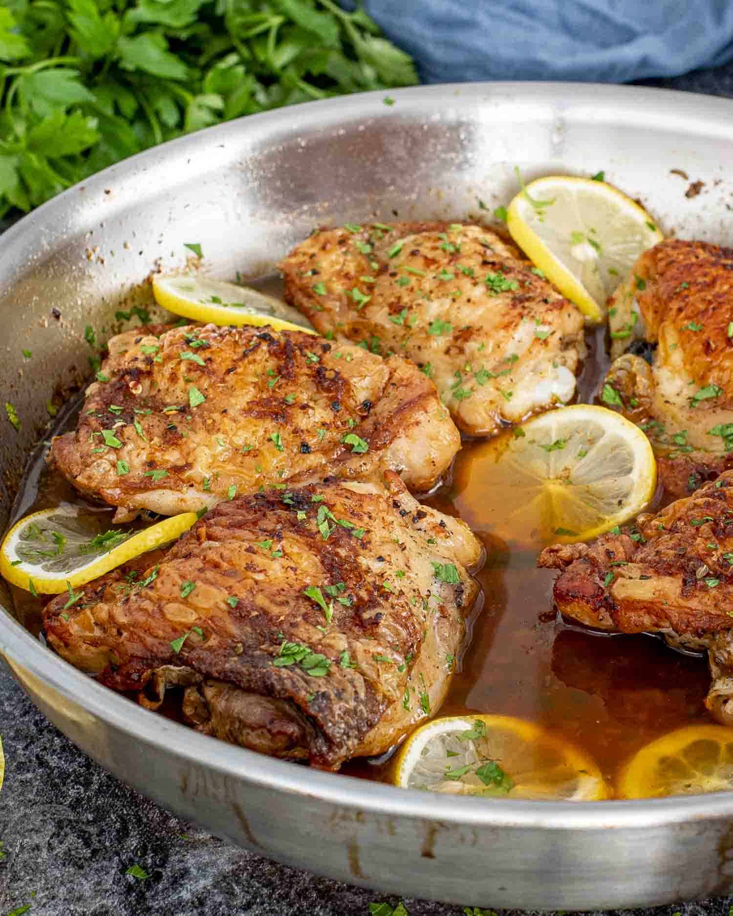 crispy lemon chicken thighs fresh out of the oven in a skillet garnished with parsley and lemon slices.