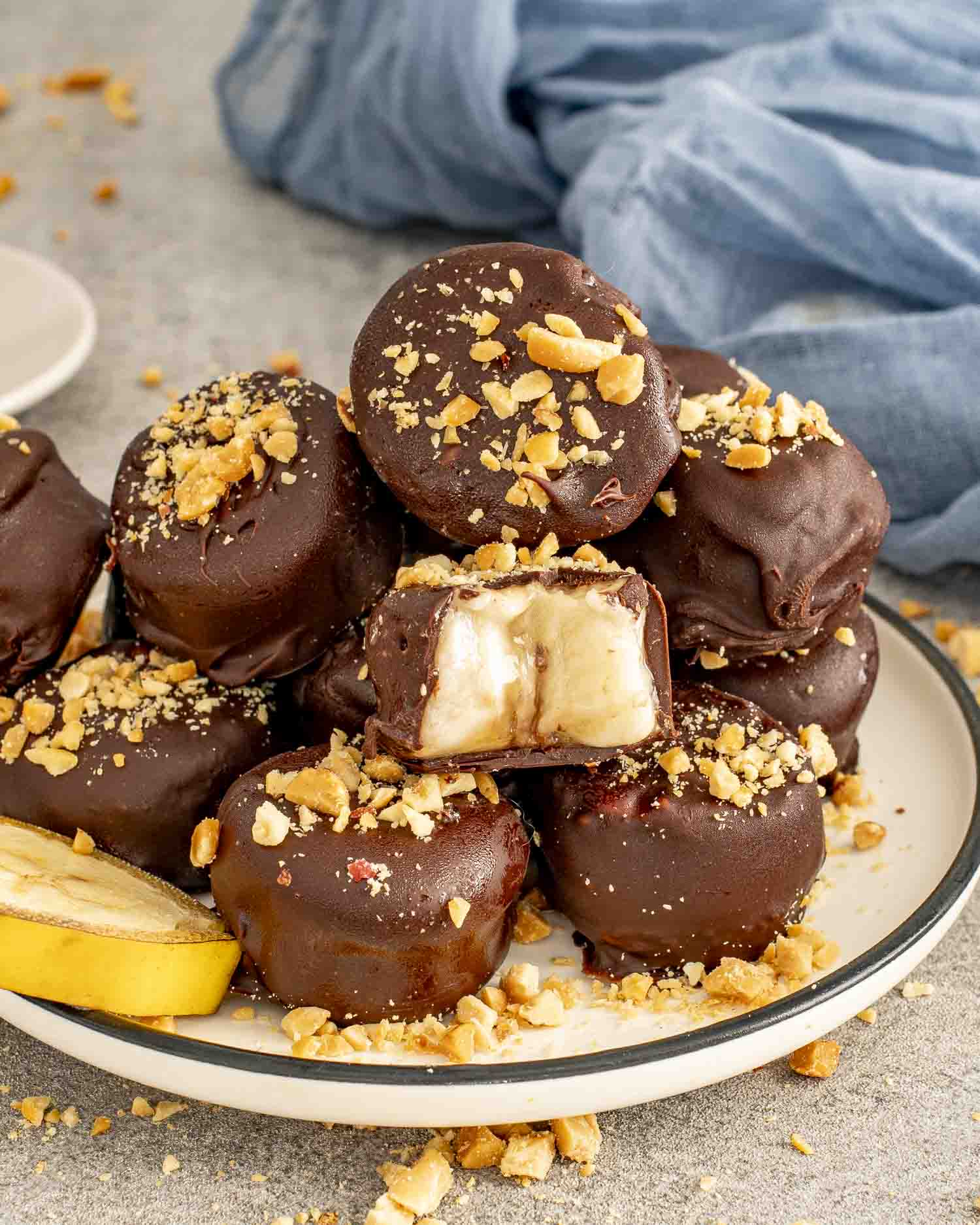 Stack of frozen chocolate-covered banana bites sprinkled with crushed nuts, one showing creamy banana center on a plate.