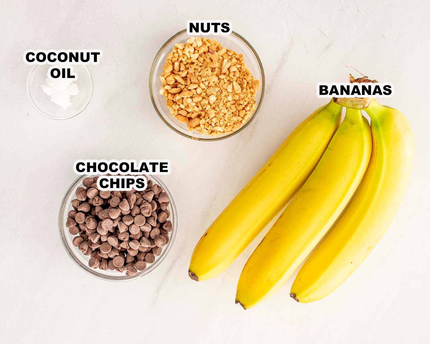 Overhead view of ripe bananas, chocolate chips, coconut oil, and chopped nuts on a white surface for banana bites.