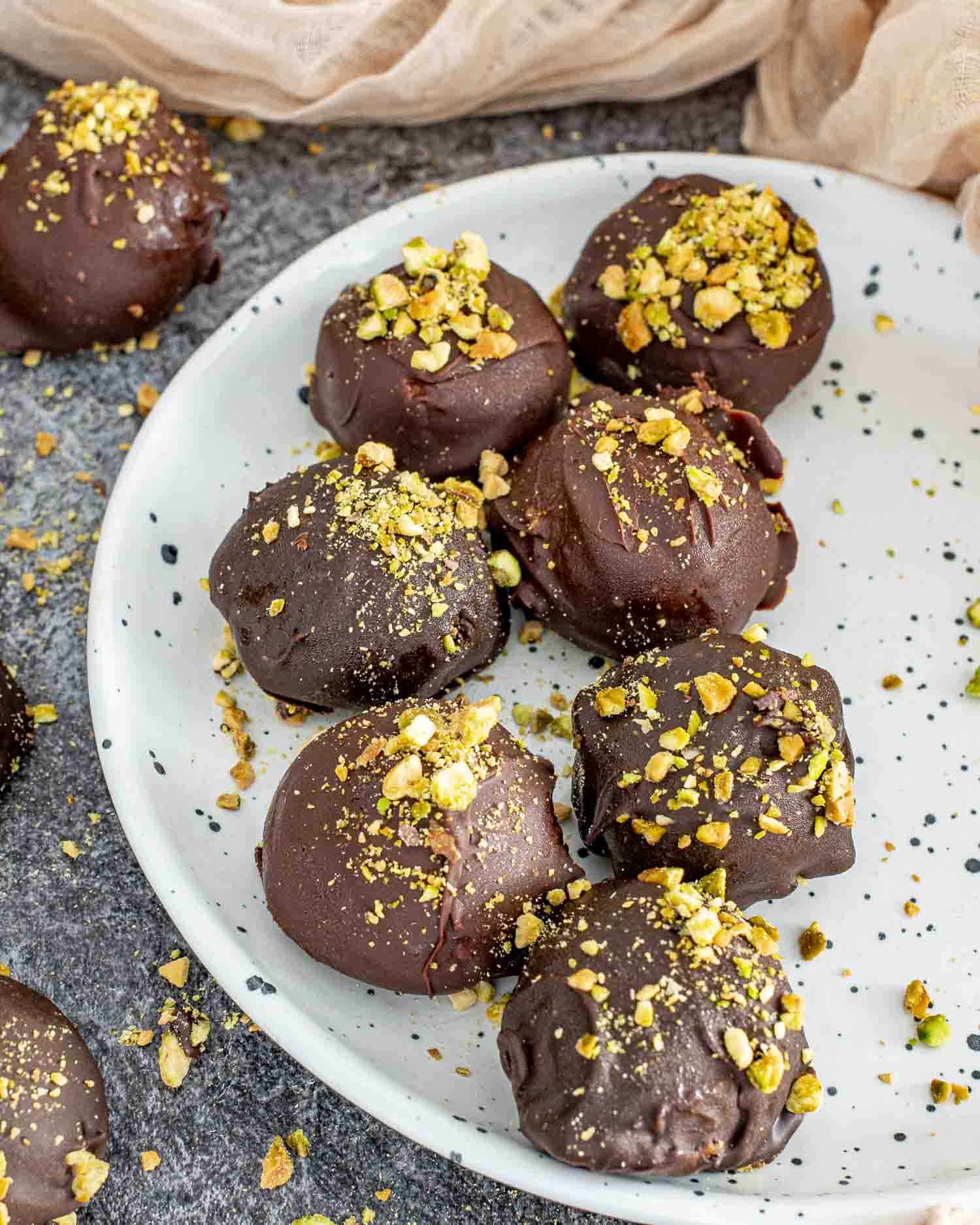 A white speckled plate filled with Frozen Pistachio and Date Chocolate Bites, coated in dark chocolate and topped with crushed pistachios. One bite has a small piece broken off, revealing a creamy, nutty pistachio and date filling. Pistachio crumbs are scattered on the plate and surrounding dark textured surface, with soft beige fabric in the background.