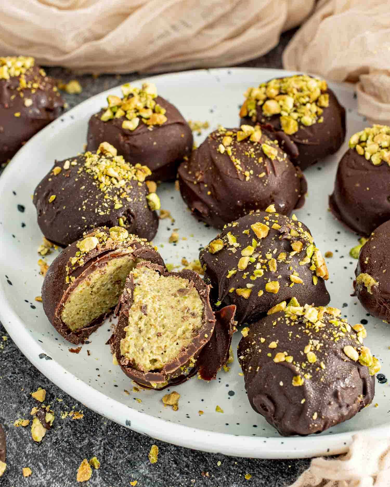 A white speckled plate filled with Frozen Pistachio and Date Chocolate Bites, coated in dark chocolate and sprinkled with crushed pistachios. One bite is cut open, revealing a creamy, nutty pistachio and date filling. Pistachio crumbs are scattered around the plate, set on a dark textured surface with soft beige fabric in the background.