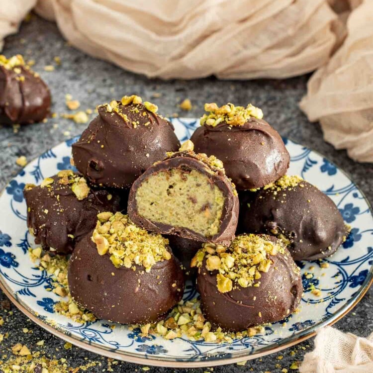 A plate of Frozen Pistachio and Date Chocolate Bites, coated in a glossy dark chocolate shell and topped with crushed pistachios. One bite is cut in half, revealing the creamy, nutty pistachio and date filling inside. The plate is set on a textured dark surface with scattered pistachio crumbs around it, with soft beige fabric in the background adding warmth to the scene.