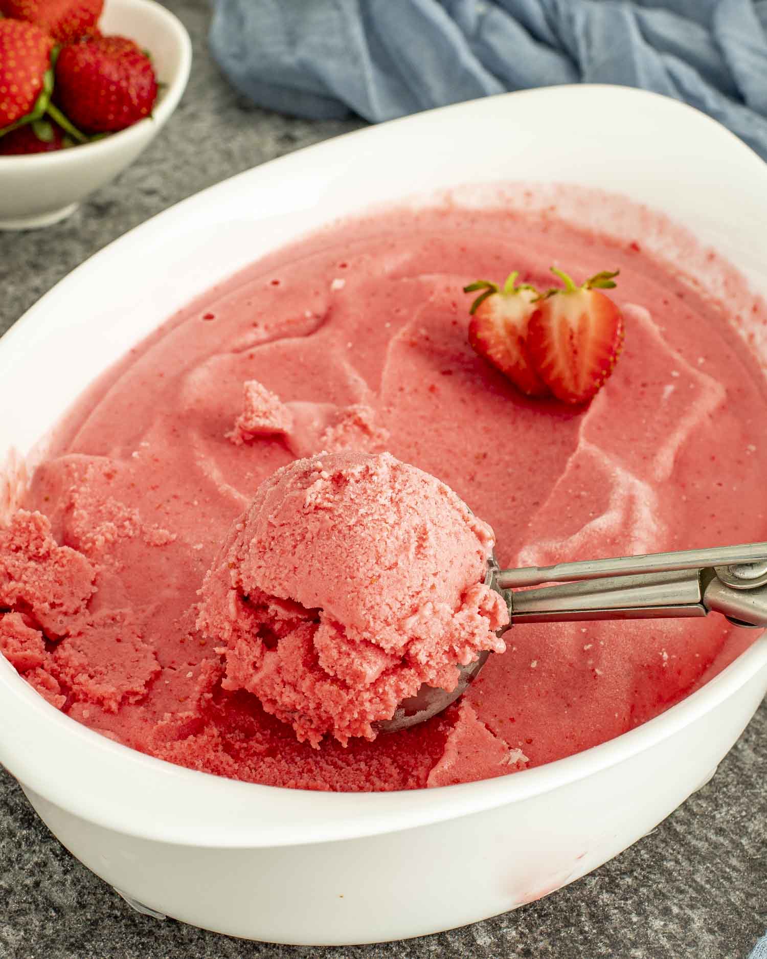 A white dish filled with frozen strawberry yogurt, one scoop served with a scooper, garnished with fresh berries.