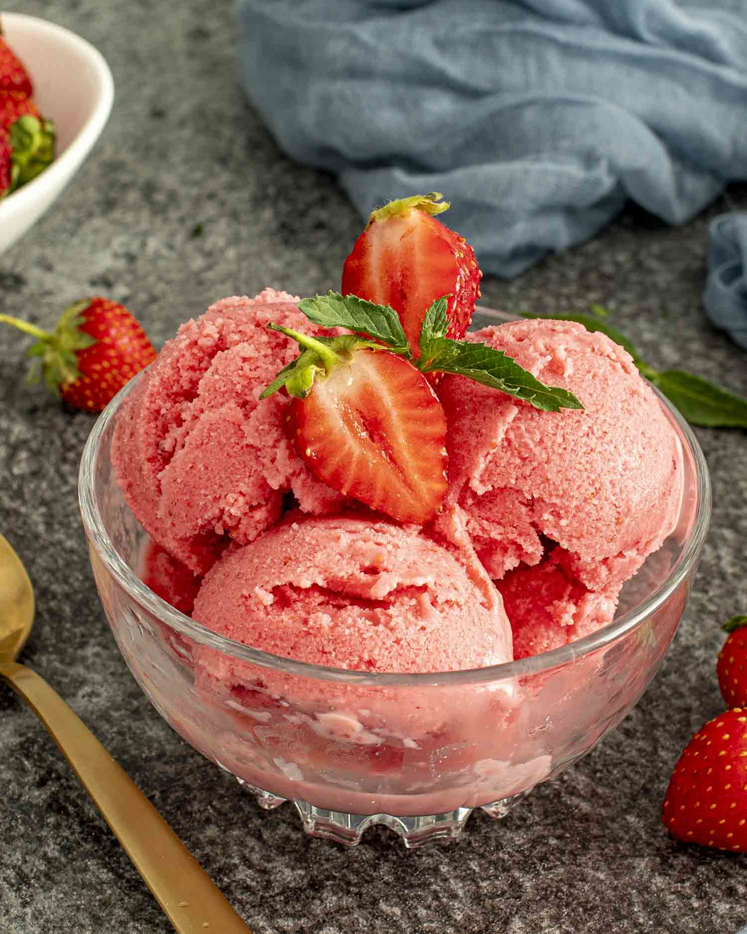 Strawberry yogurt scoops in a bowl, styled with halved strawberries and mint, set on a dark countertop.