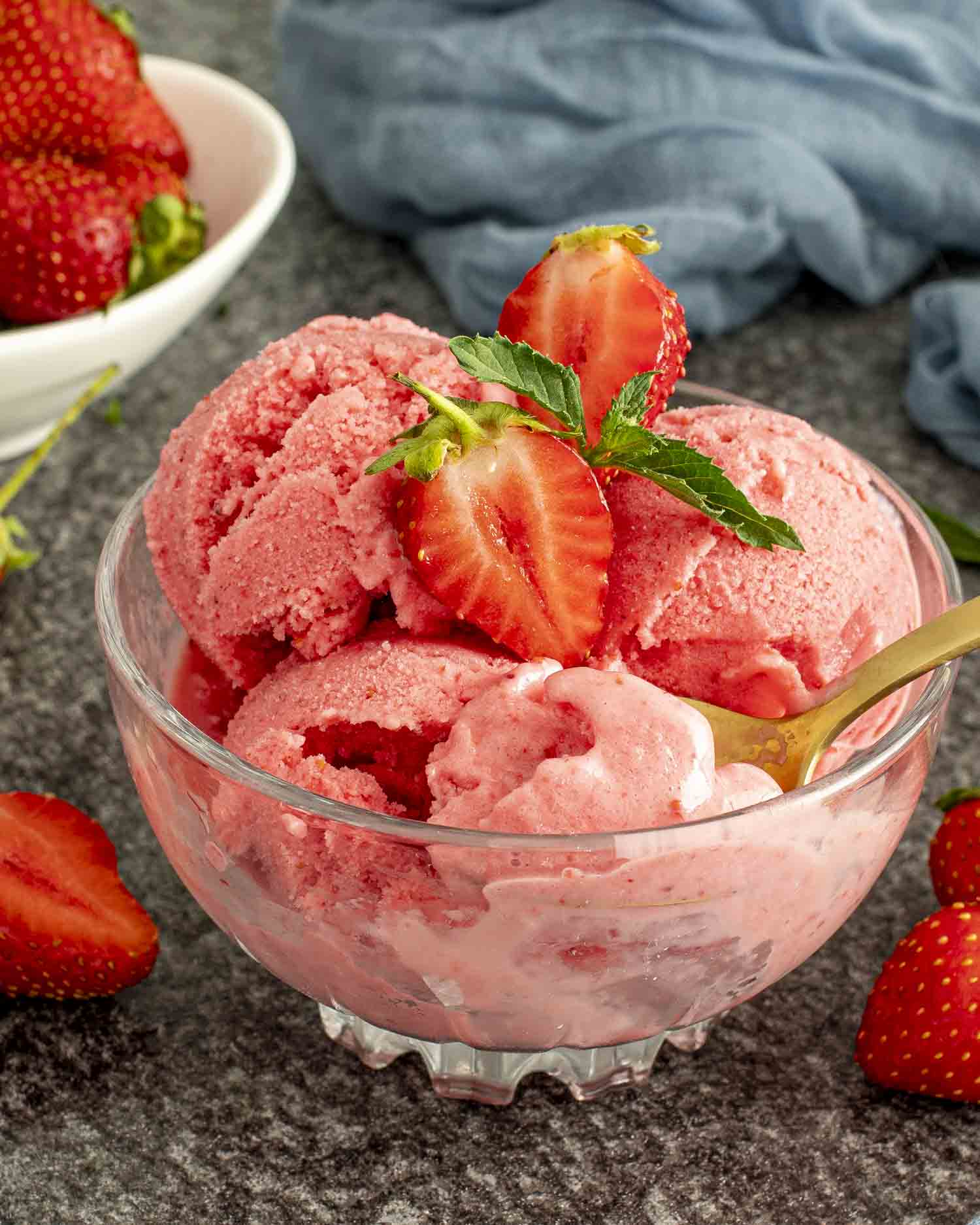 Scoops of frozen strawberry yogurt in a glass bowl topped with fresh strawberries and mint, golden spoon inside.