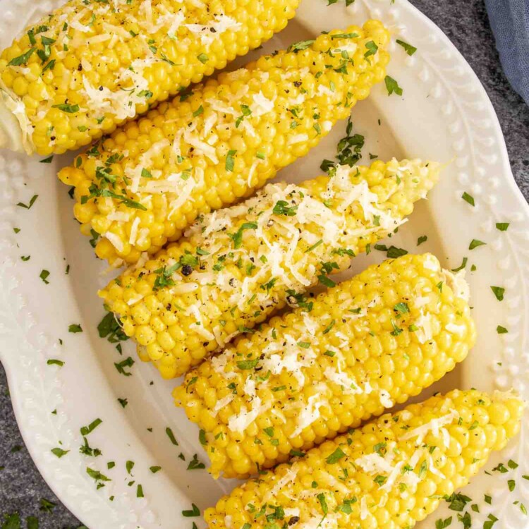 Overhead view of five corn cobs smothered in garlic butter, Parmesan, and parsley on a white serving dish.