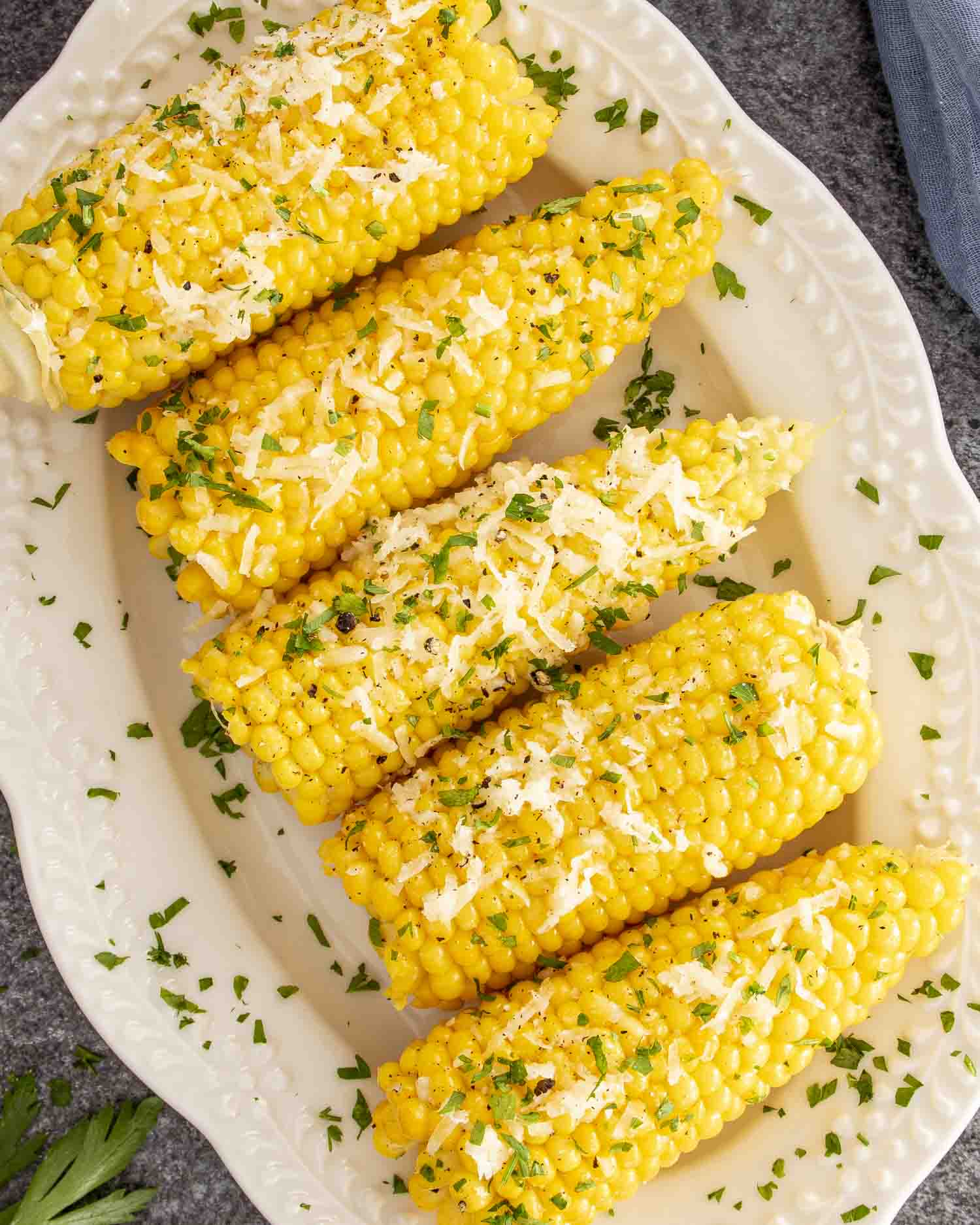 Overhead view of five corn cobs smothered in garlic butter, Parmesan, and parsley on a white serving dish.