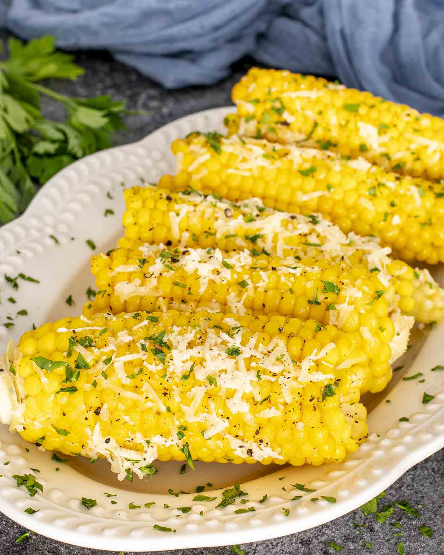 Four garlic Parmesan corn cobs arranged on a white platter, garnished with chopped parsley and shredded cheese, perfect for summer.
