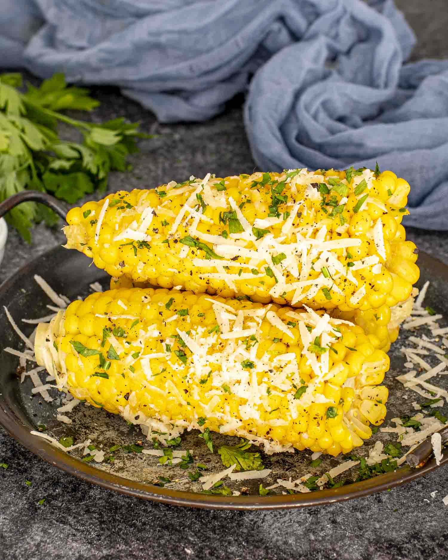 Close-up of two buttery corn cobs sprinkled with Parmesan and fresh parsley, displayed on a dark plate with herbs.