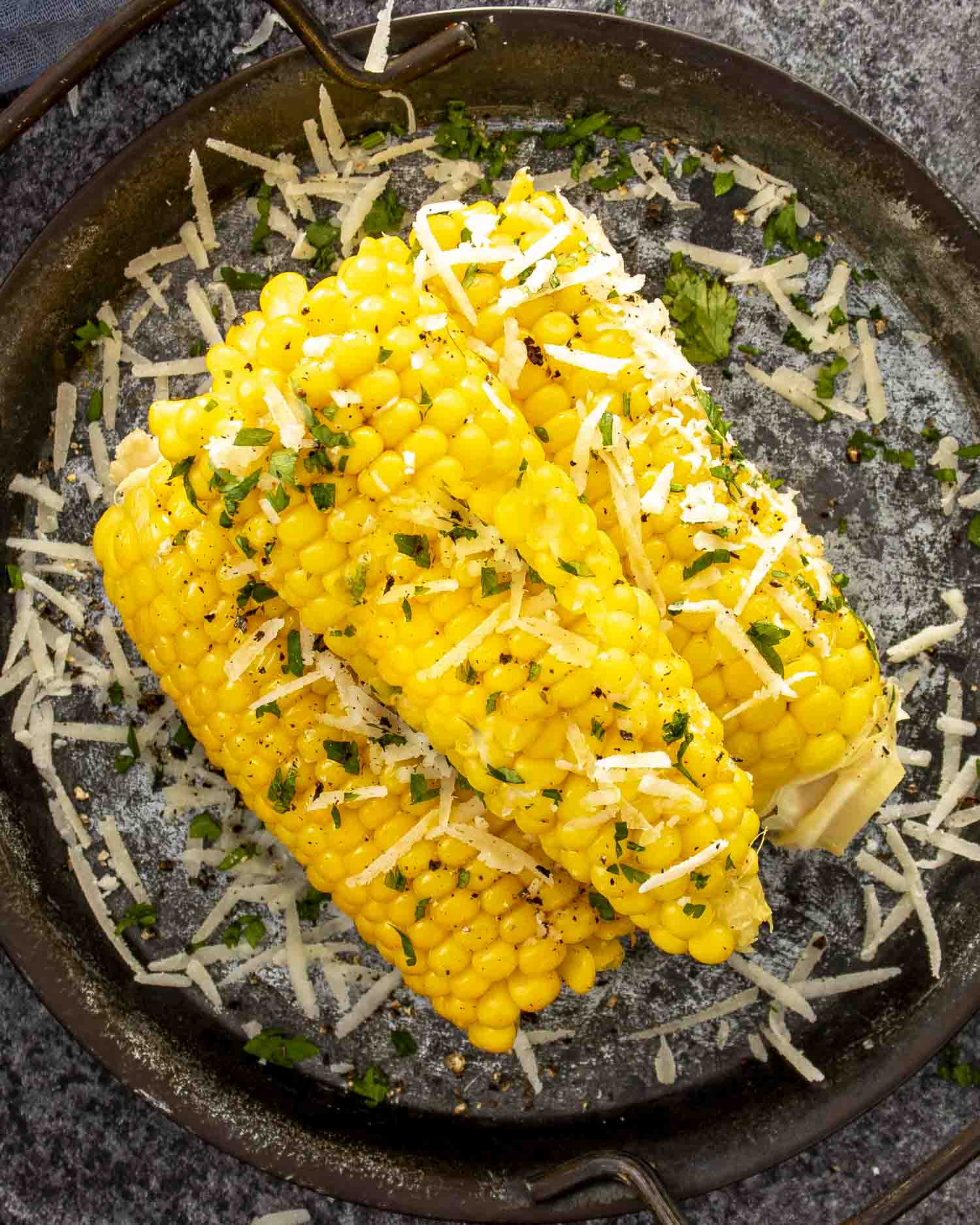 Two ears of corn on the cob topped with Parmesan cheese and parsley, served in a rustic metal pan.