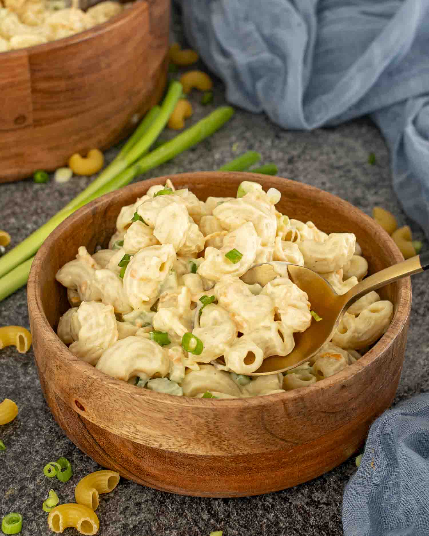 Hawaiian macaroni salad served in a wooden bowl with a spoon, garnished with green onions, placed on gray surface.
