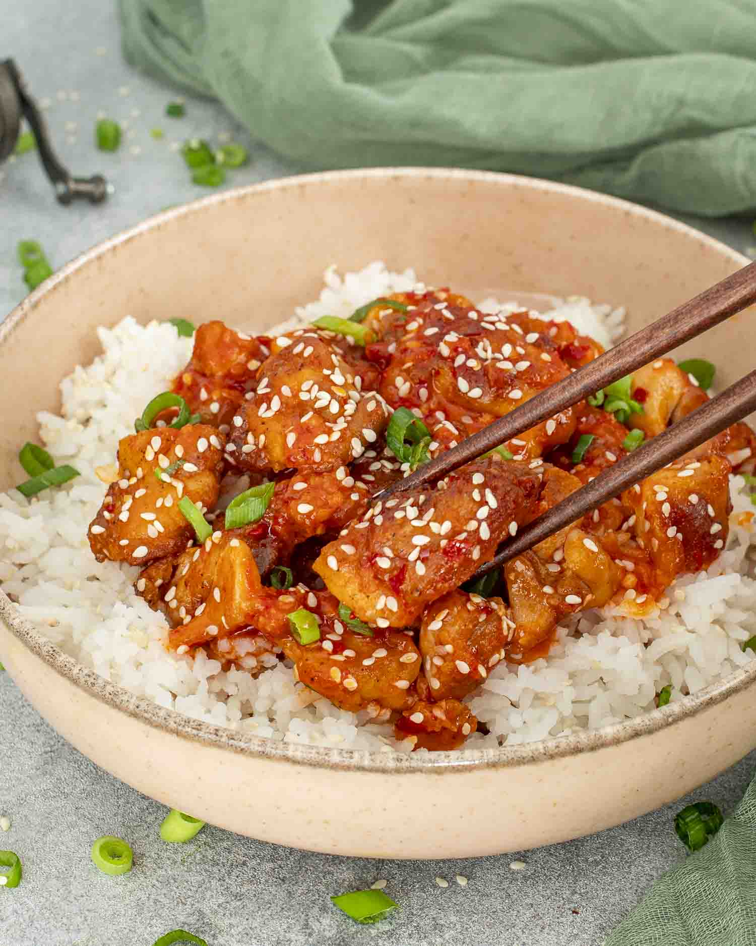 Chopsticks picking up saucy Korean BBQ chicken from a bowl of white rice, topped with sesame seeds.