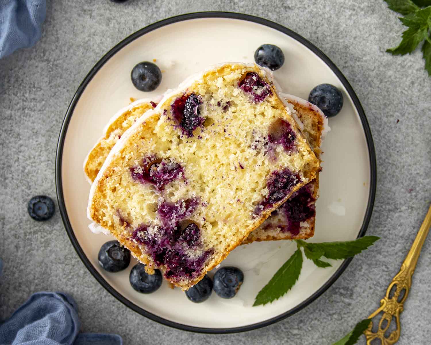 Top view of moist lemon blueberry bread slices on a plate surrounded by fresh blueberries and mint leaves.