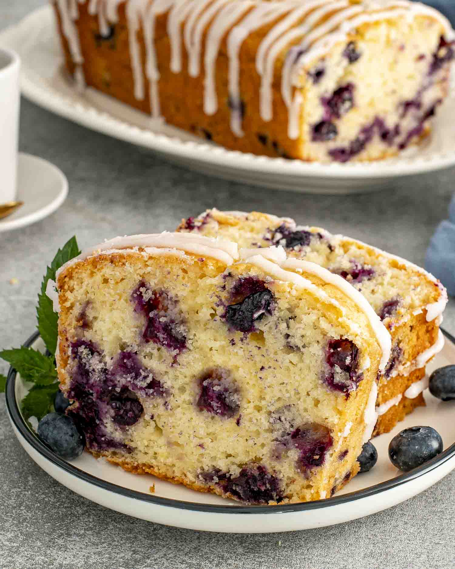 Two slices of lemon blueberry bread with sweet glaze drizzle, bursting with fresh blueberries and golden edges on a white plate.