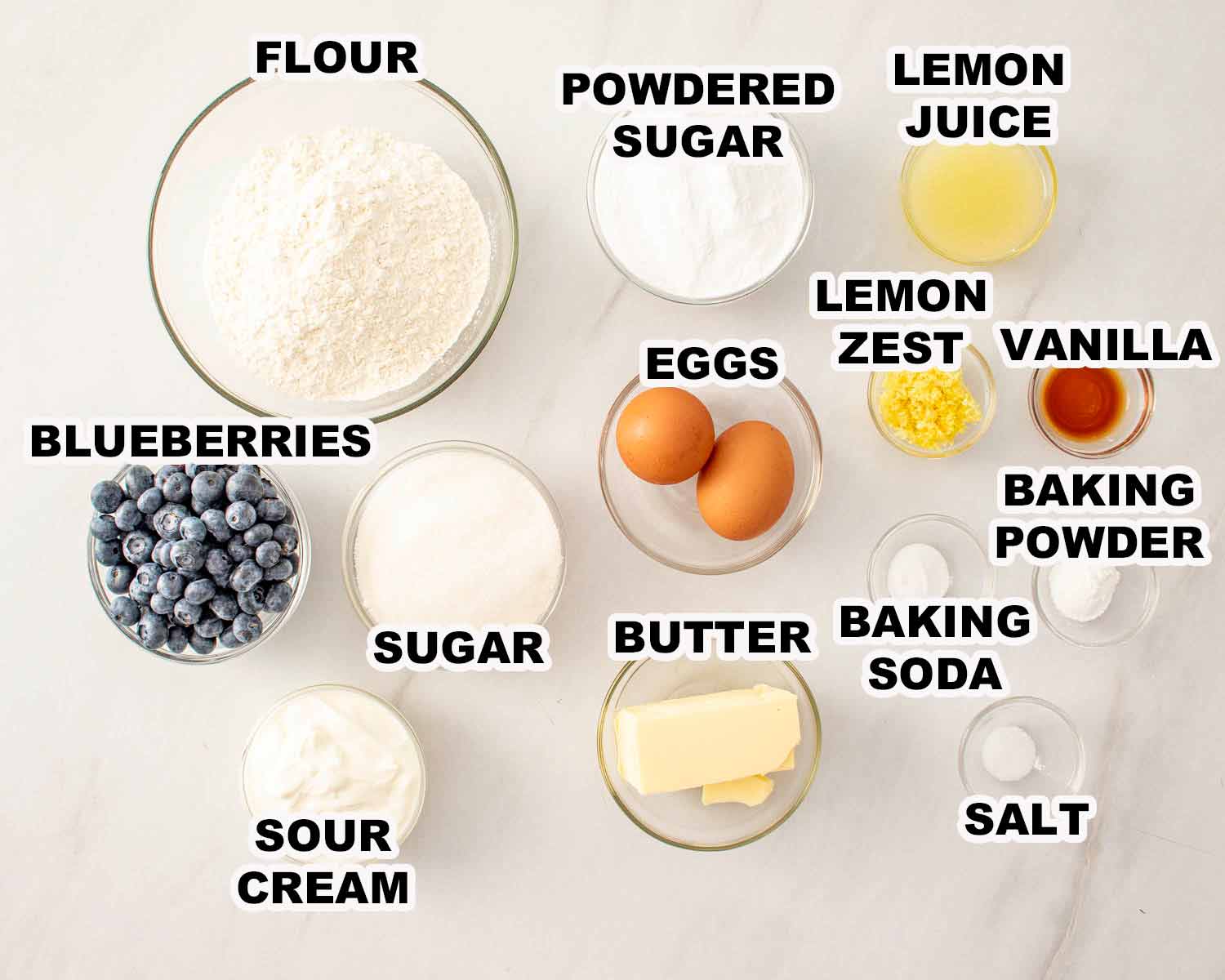All ingredients for lemon blueberry bread including flour, blueberries, eggs, butter, sugar, sour cream, lemon, and vanilla arranged neatly.