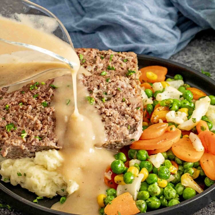 plate of meatloaf slices served with mashed potatoes and mixed vegetables, topped with creamy gravy being poured from a glass pitcher.