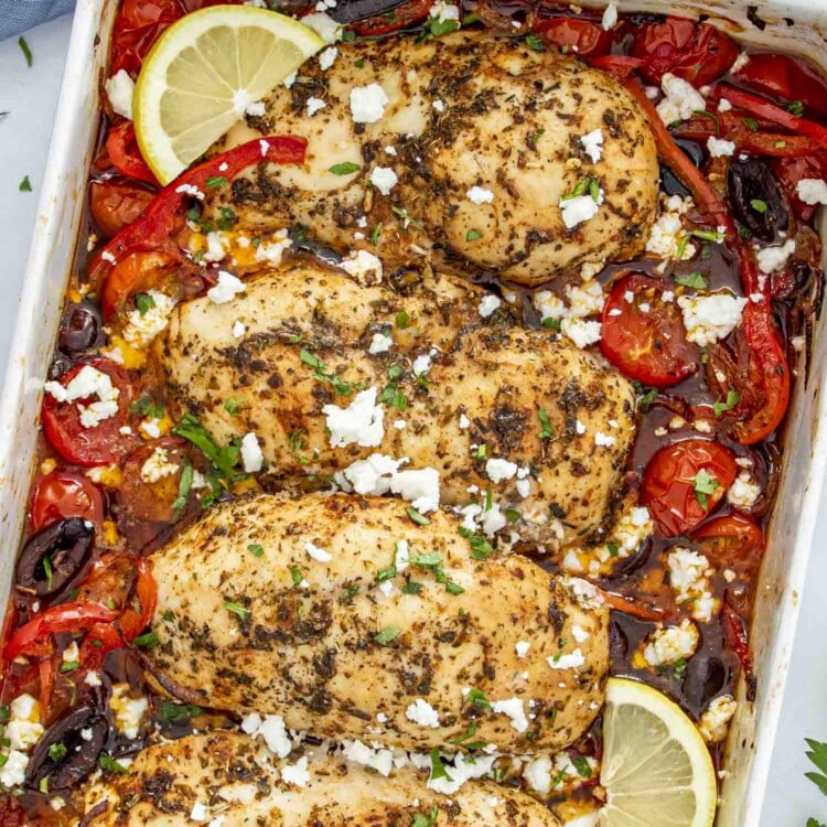 A close-up of Mediterranean Chicken Bake featuring golden-brown chicken breasts seasoned with herbs, nestled among roasted cherry tomatoes, red bell peppers, Kalamata olives, and red onions.