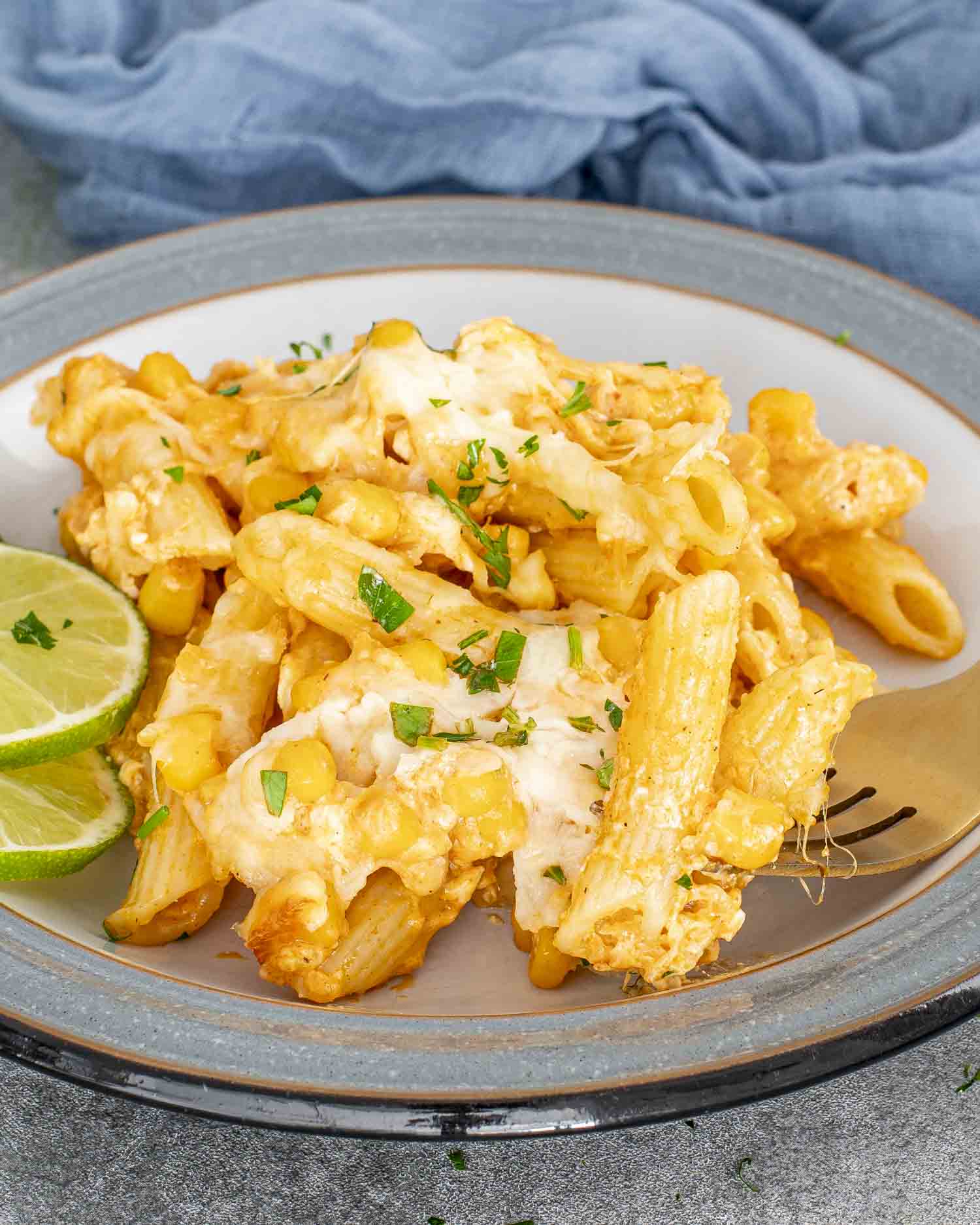 Plate of cheesy Mexican Street Corn Pasta Bake served with fresh lime slices and a gold fork, garnished with chopped parsley.