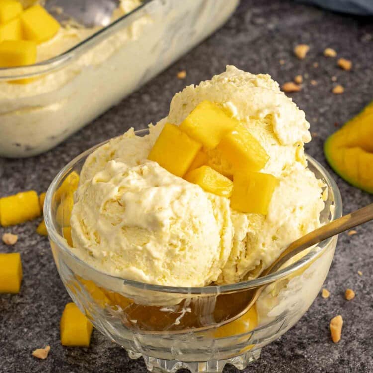Close-up of no churn mango ice cream in a glass dish, garnished with mango chunks and a gold spoon.